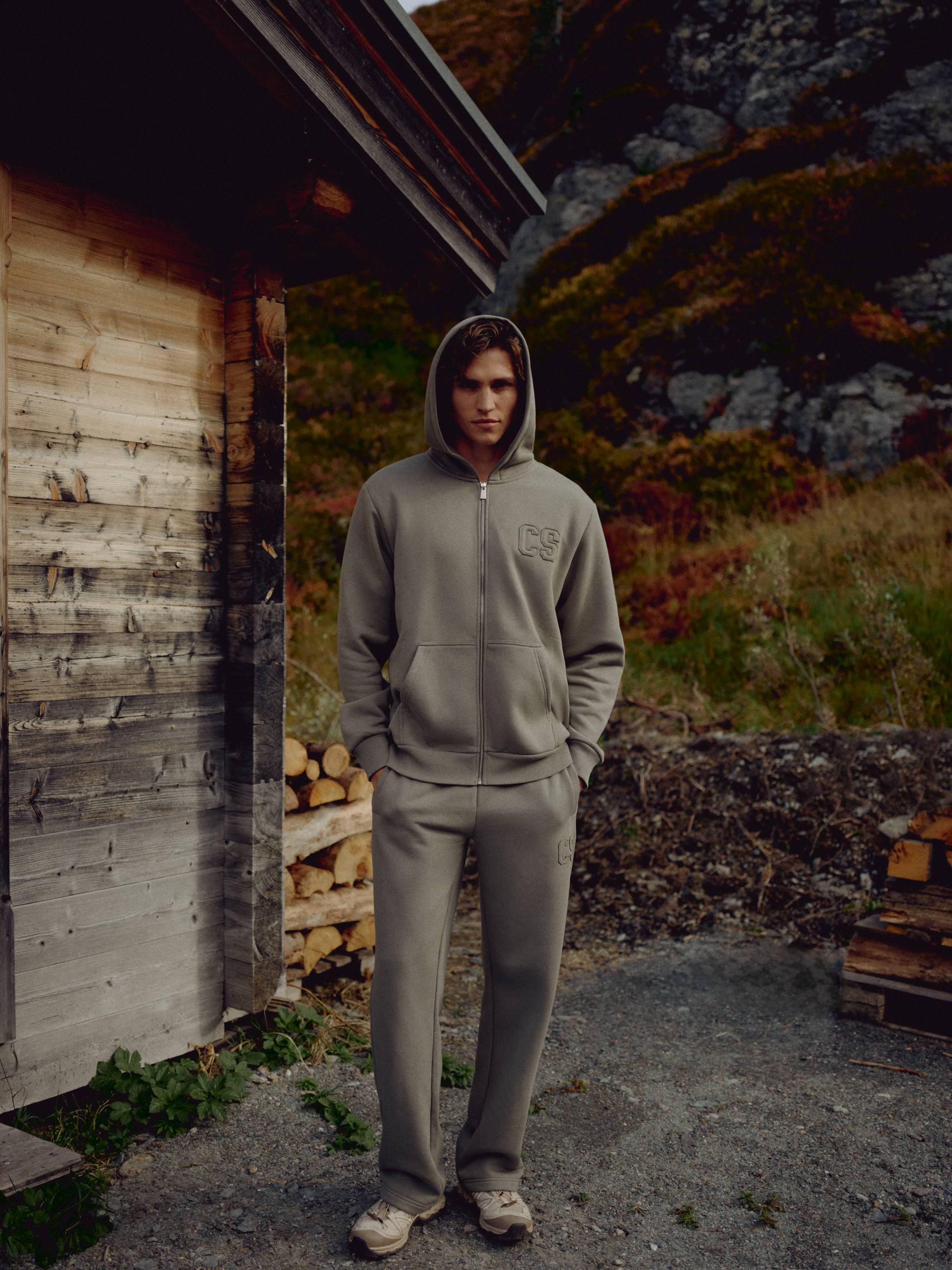 Man wearing a khaki CASPARA CS zip hoodie standing next to a rustic wooden cabin.