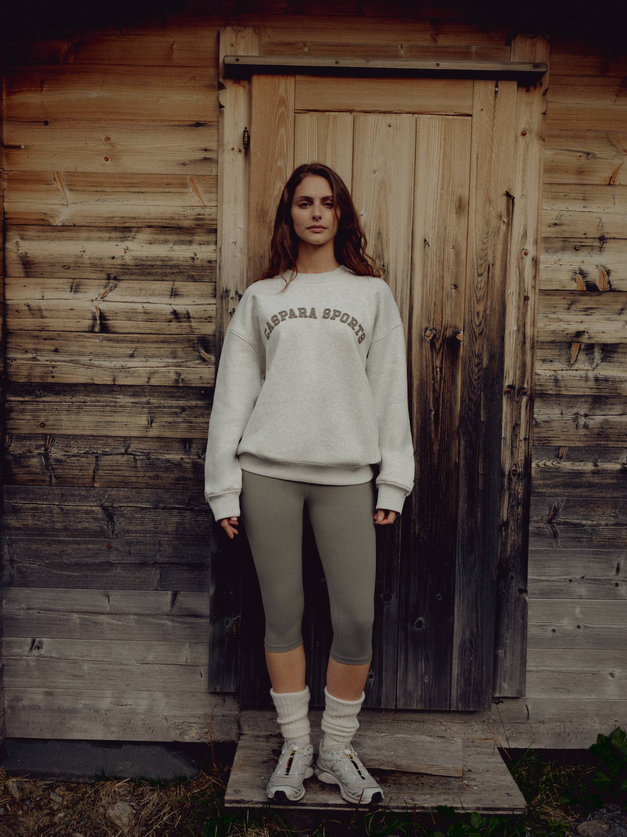 Woman wearing a grey melange CASPARA CS crewneck sweatshirt in front of a rustic wooden door.