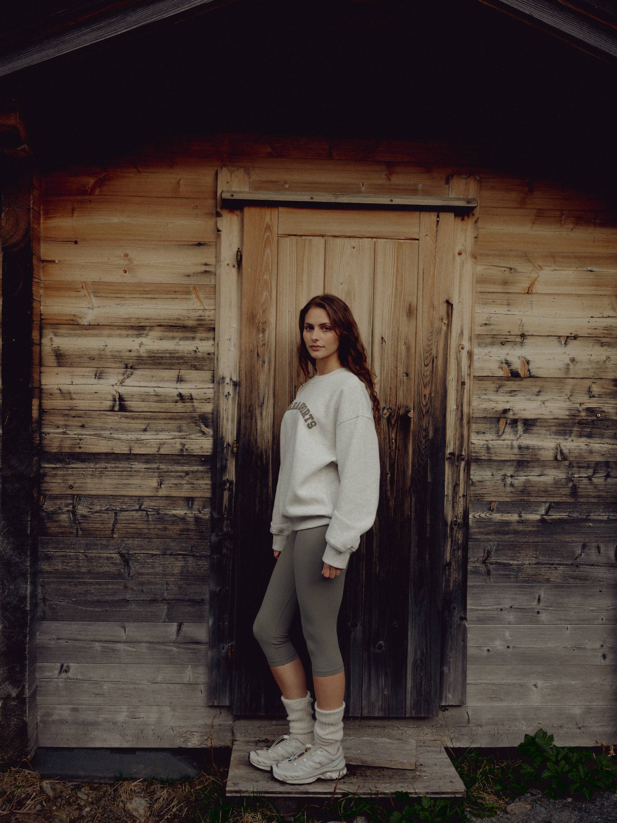 Woman wearing a grey melange CASPARA CS crewneck sweatshirt in front of a wooden cabin.