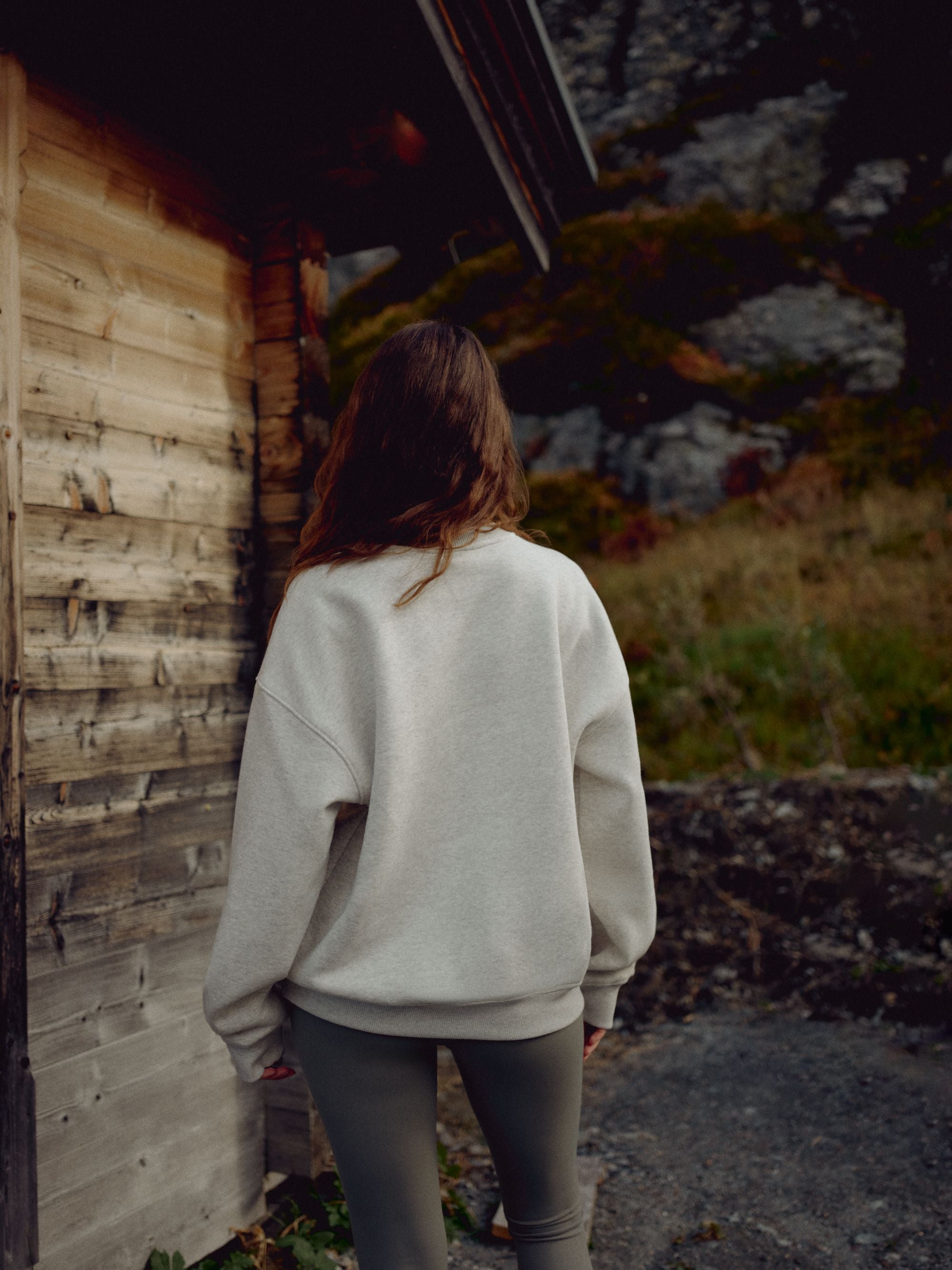 Woman wearing a grey melange CASPARA CS crewneck from behind, standing next to a wooden cabin.