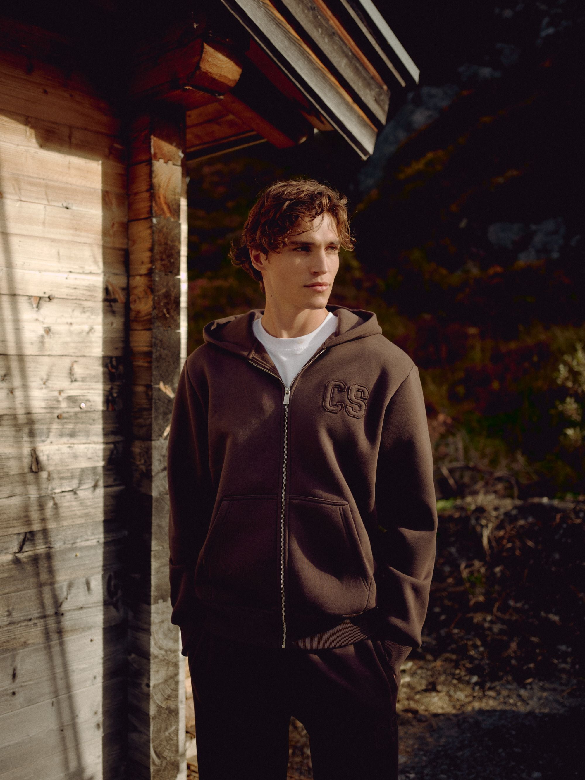 Man wearing an espresso CASPARA CS zip hoodie standing next to a wooden cabin.