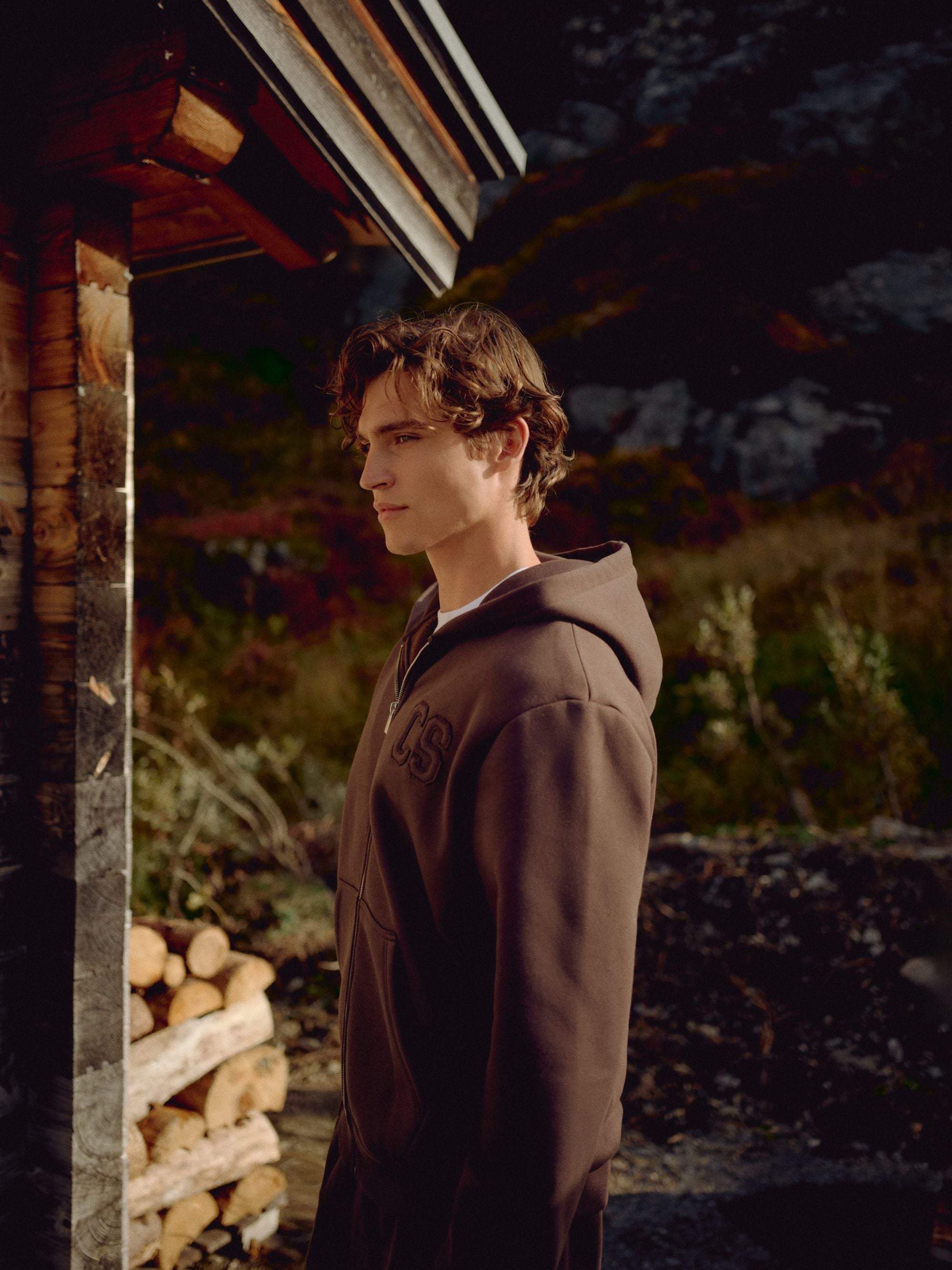 Man wearing an espresso CASPARA CS zip hoodie standing outside a wooden cabin.