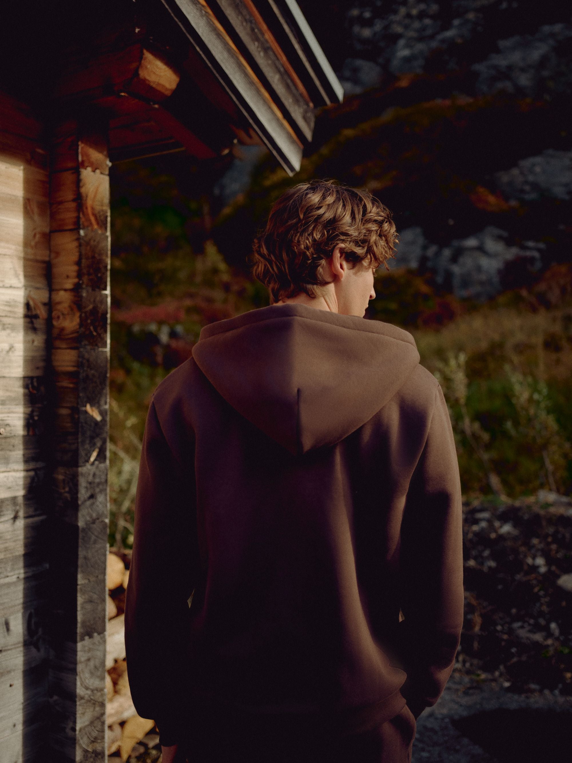 Man wearing an espresso CASPARA CS zip hoodie seen from behind by a wooden cabin.