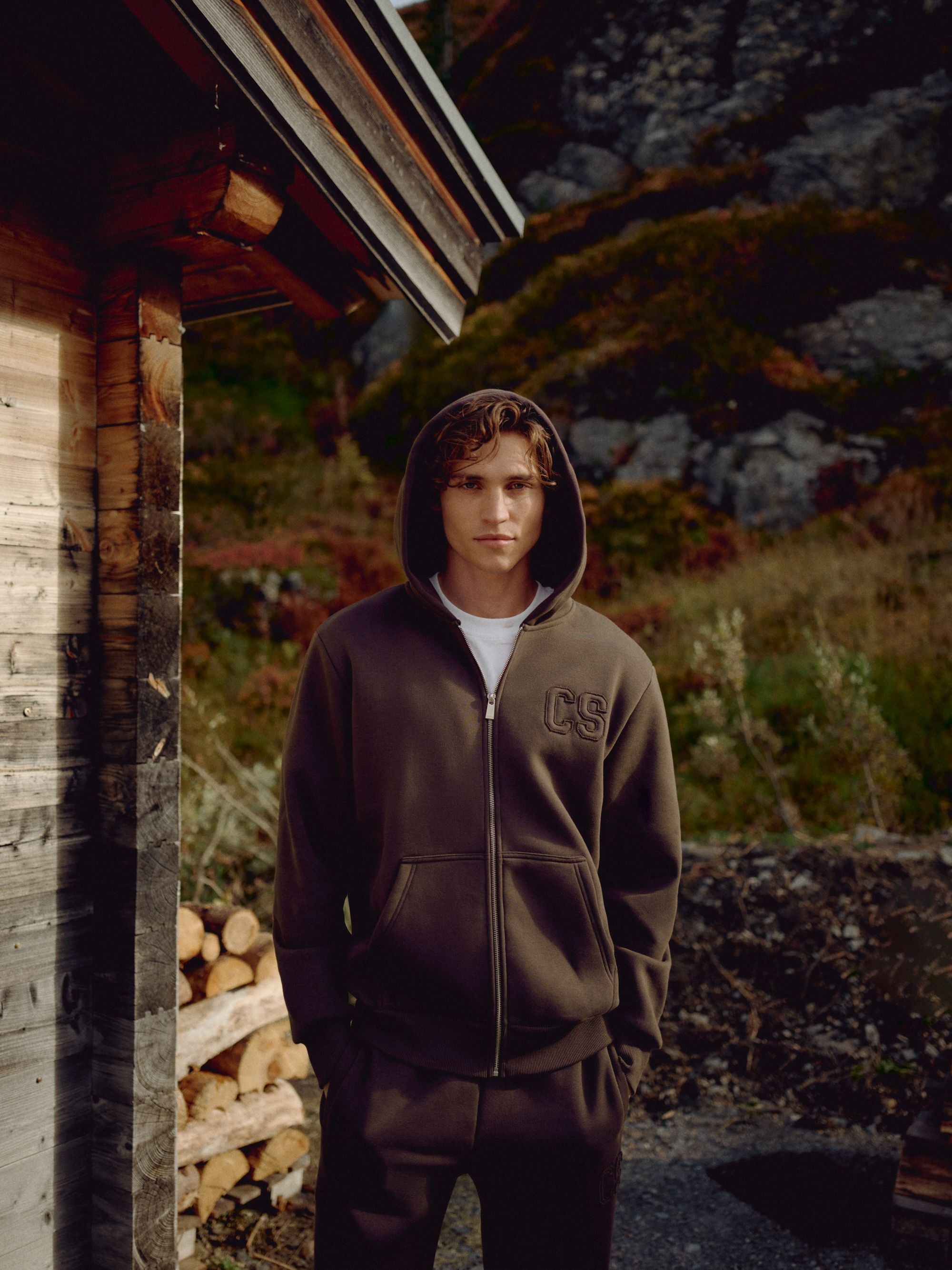 Man wearing an espresso CASPARA CS zip hoodie standing next to a wooden cabin.