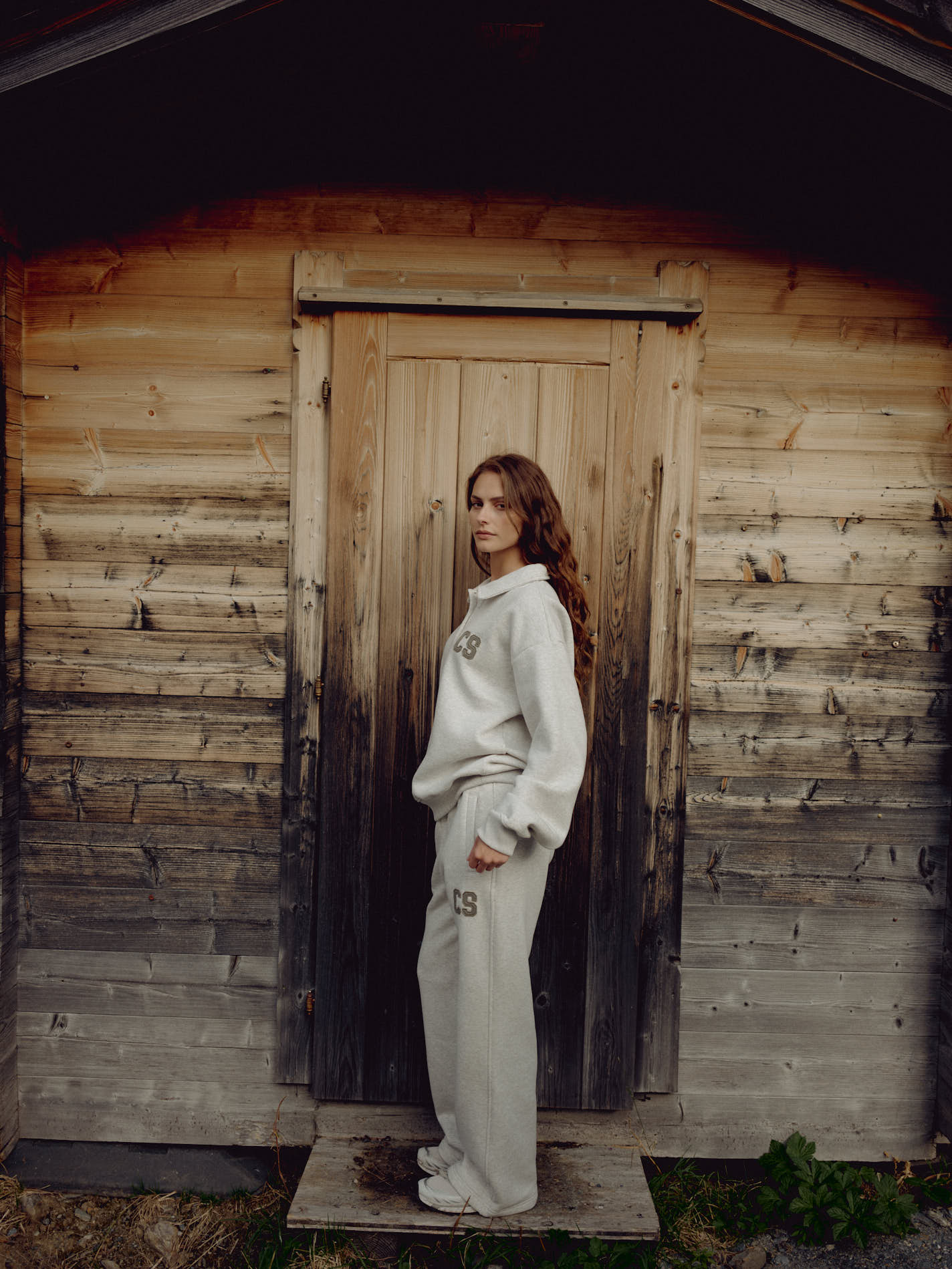 Woman wearing a grey melange CASPARA CS rugby shirt standing in front of a wooden cabin.