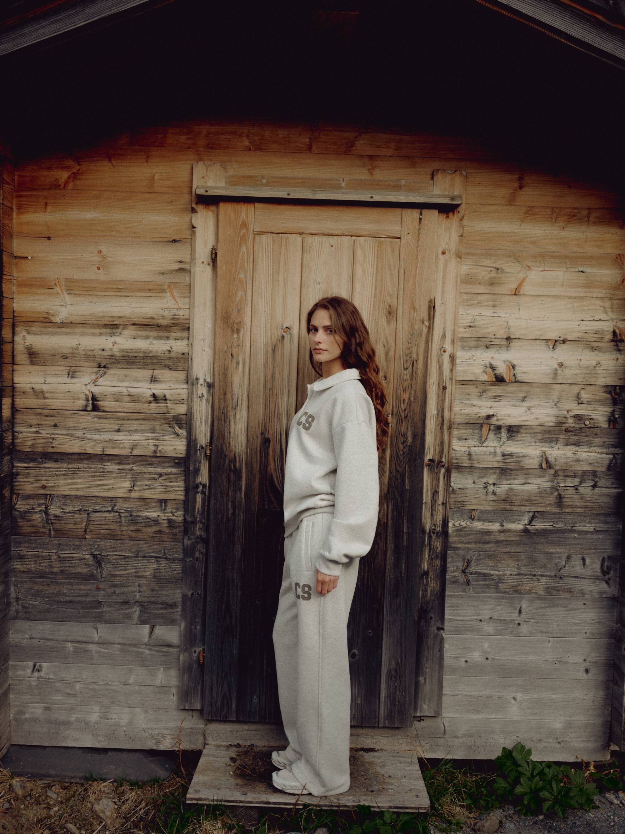 Woman wearing a grey melange CASPARA CS rugby shirt standing outside a wooden cabin.