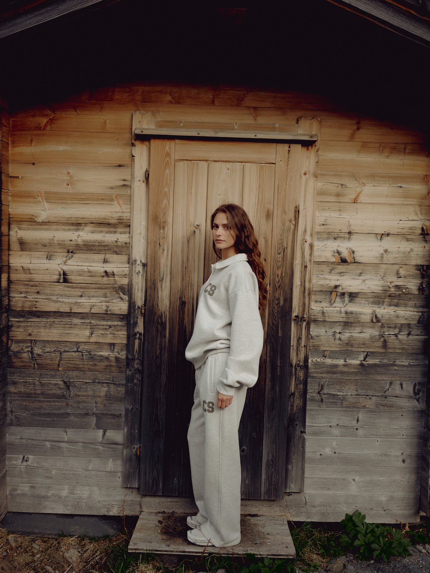 Woman wearing grey melange CASPARA CS sweatpants and sweatshirt standing in front of a wooden cabin.