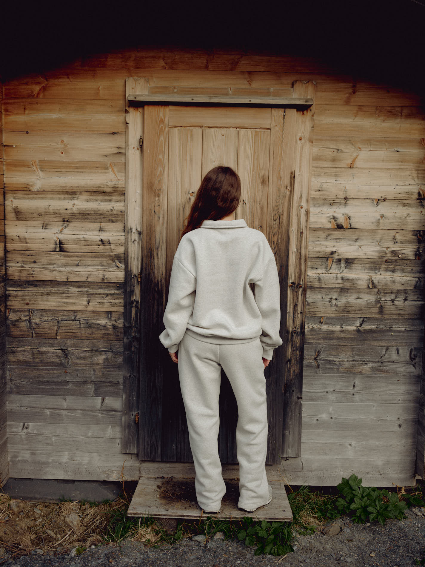 Woman wearing a grey melange CASPARA CS rugby shirt facing a rustic wooden cabin door.