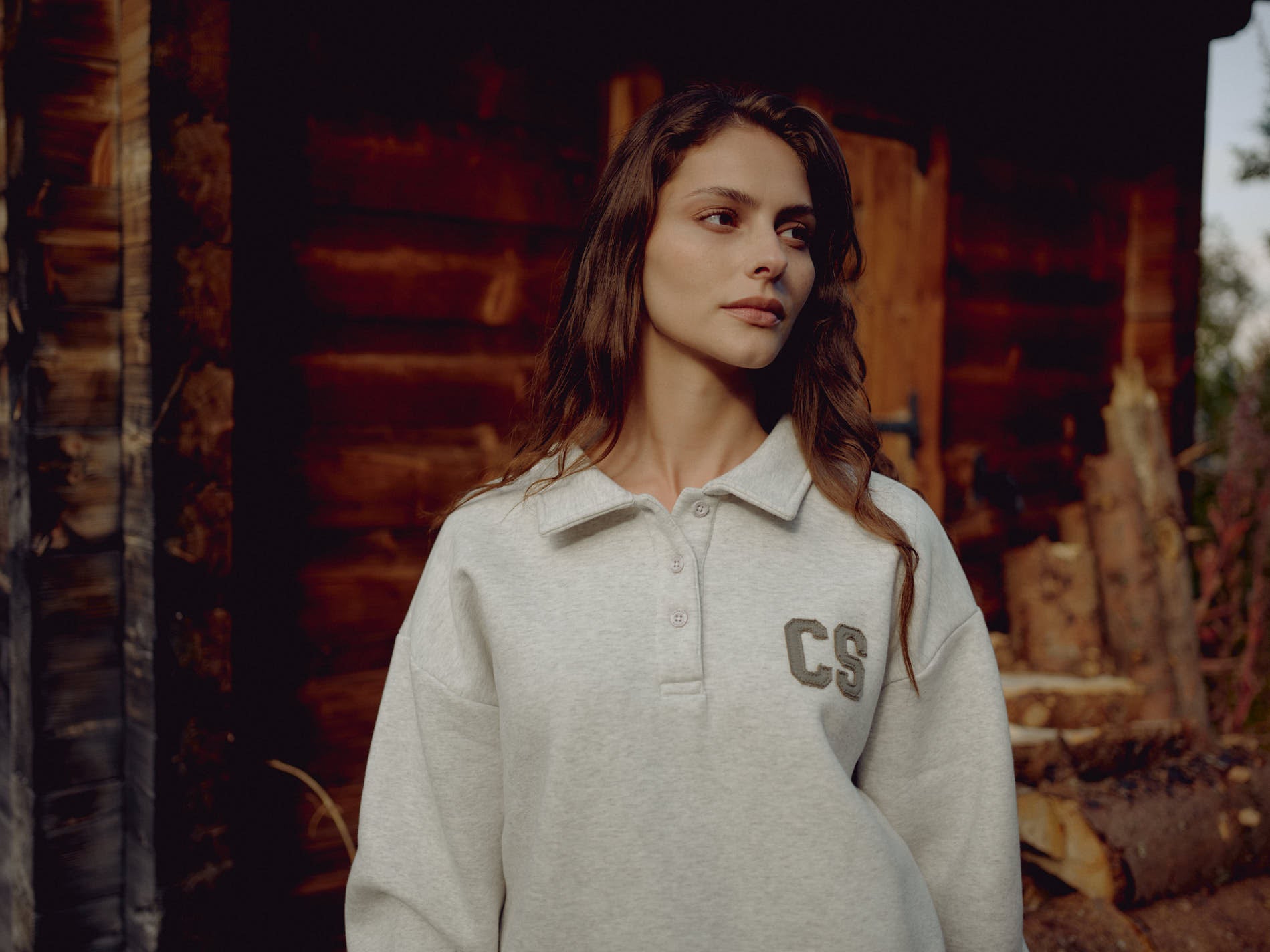 Woman wearing a grey melange CASPARA CS rugby shirt standing outside a wooden cabin.