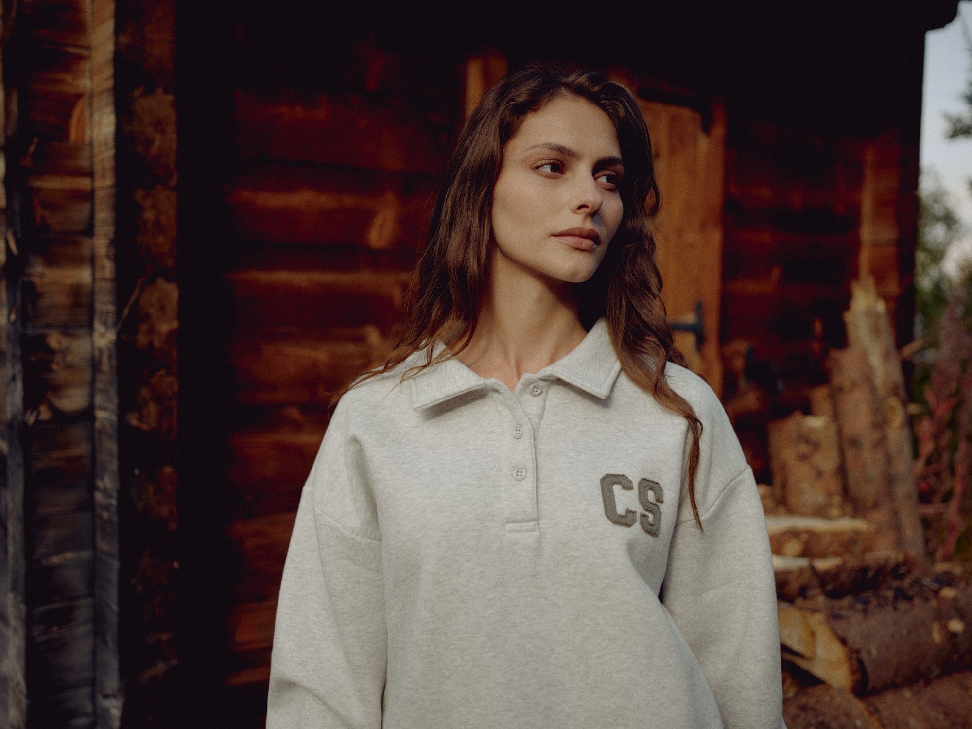 Woman wearing a grey melange CASPARA CS rugby shirt in front of a rustic wooden cabin.