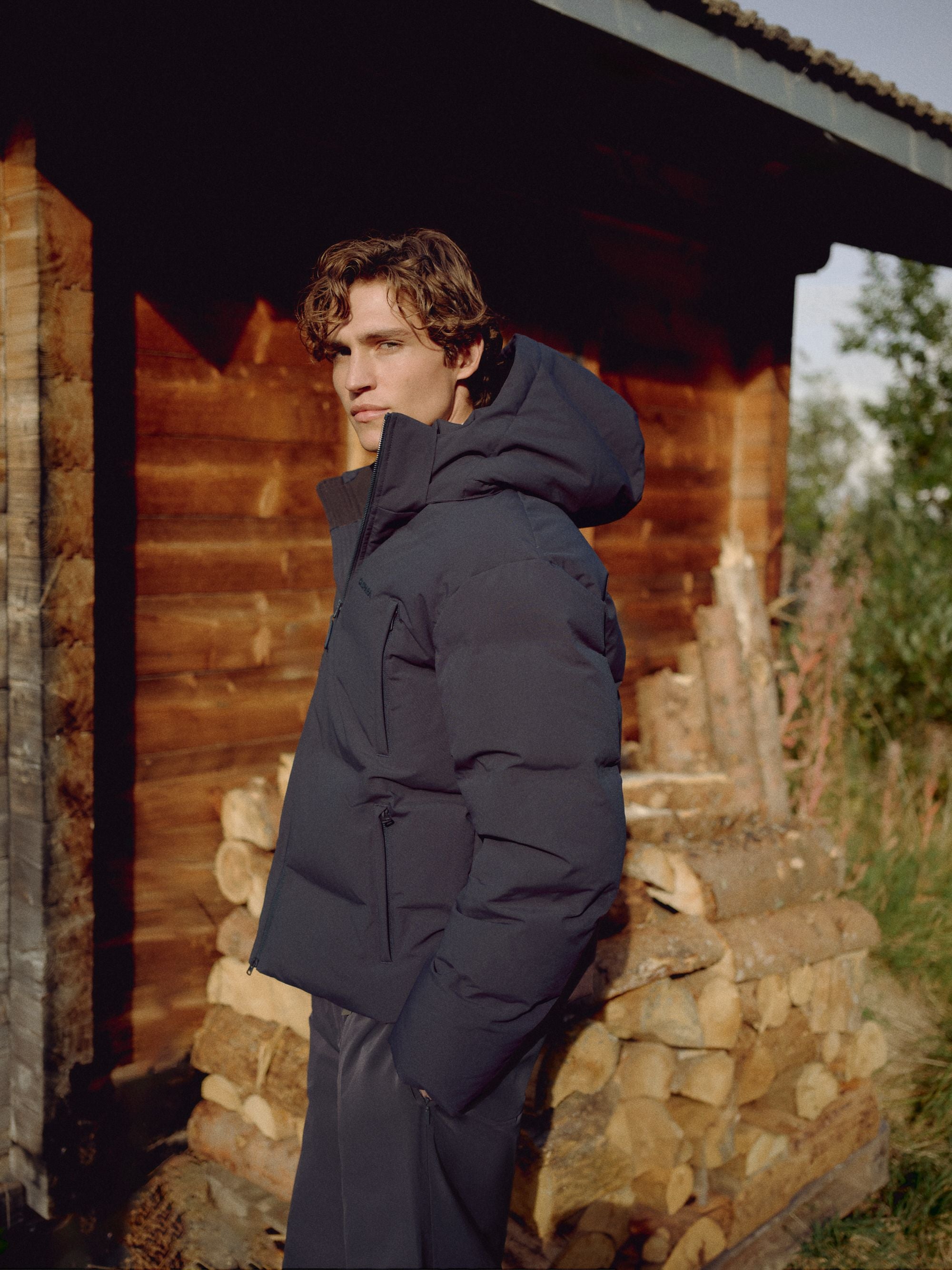 Man wearing a navy CASPARA Boreal puffer jacket outdoors beside stacked firewood.