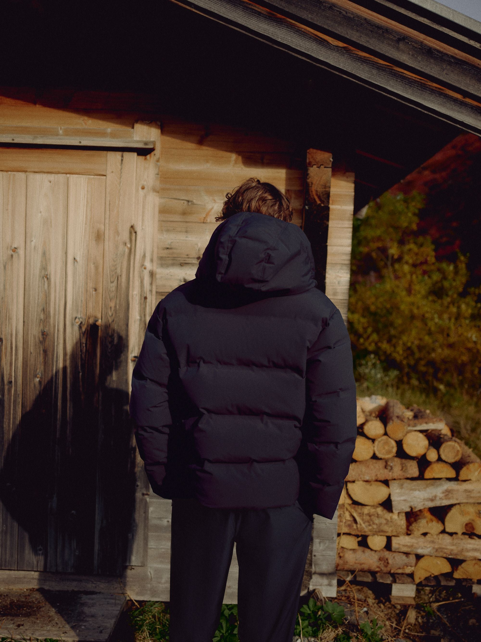 Back view of a man wearing a navy CASPARA Boreal puffer jacket by a rustic wooden cabin.