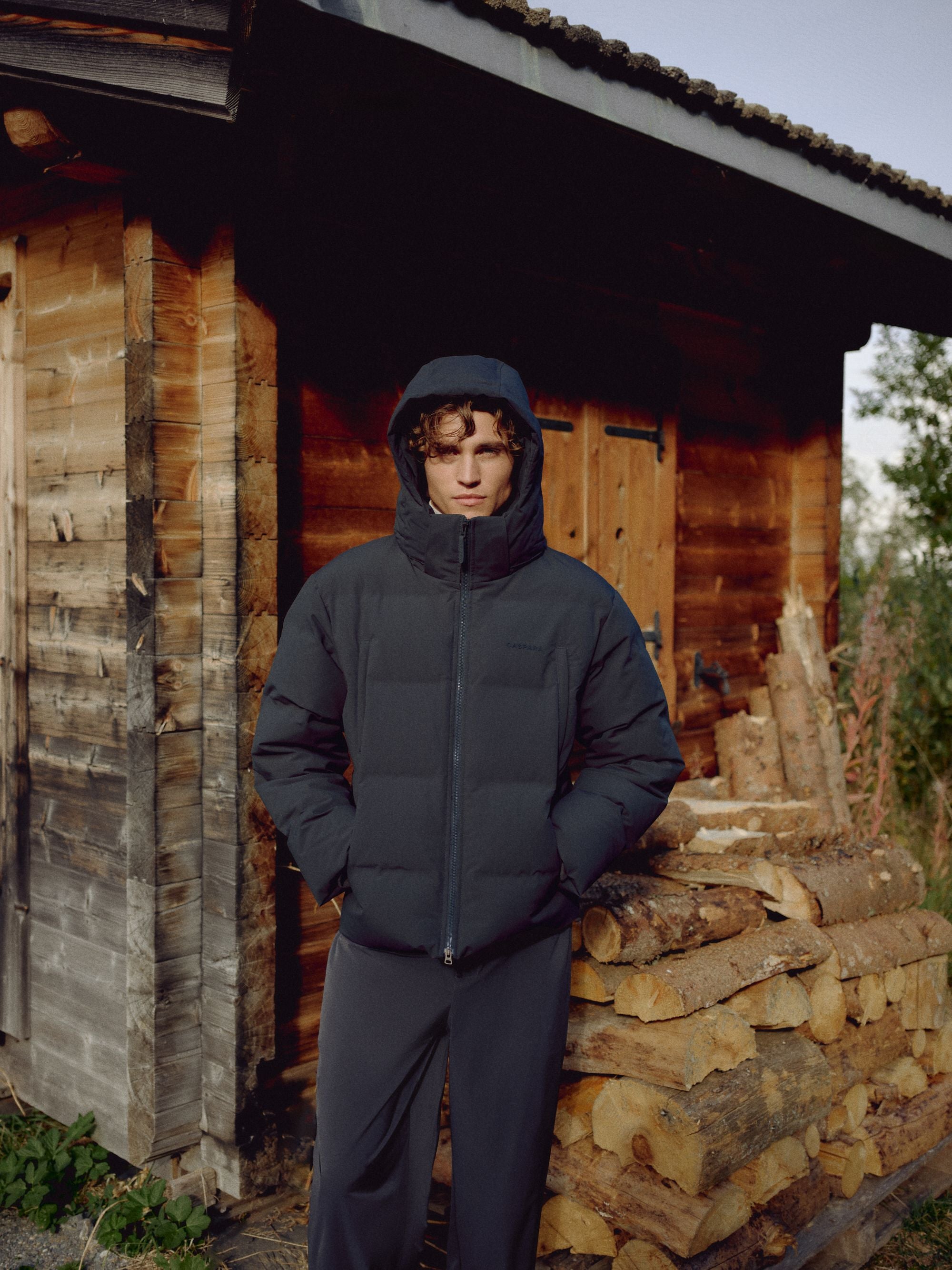 Man wearing a navy CASPARA Boreal puffer jacket with hood up outside a wooden cabin.