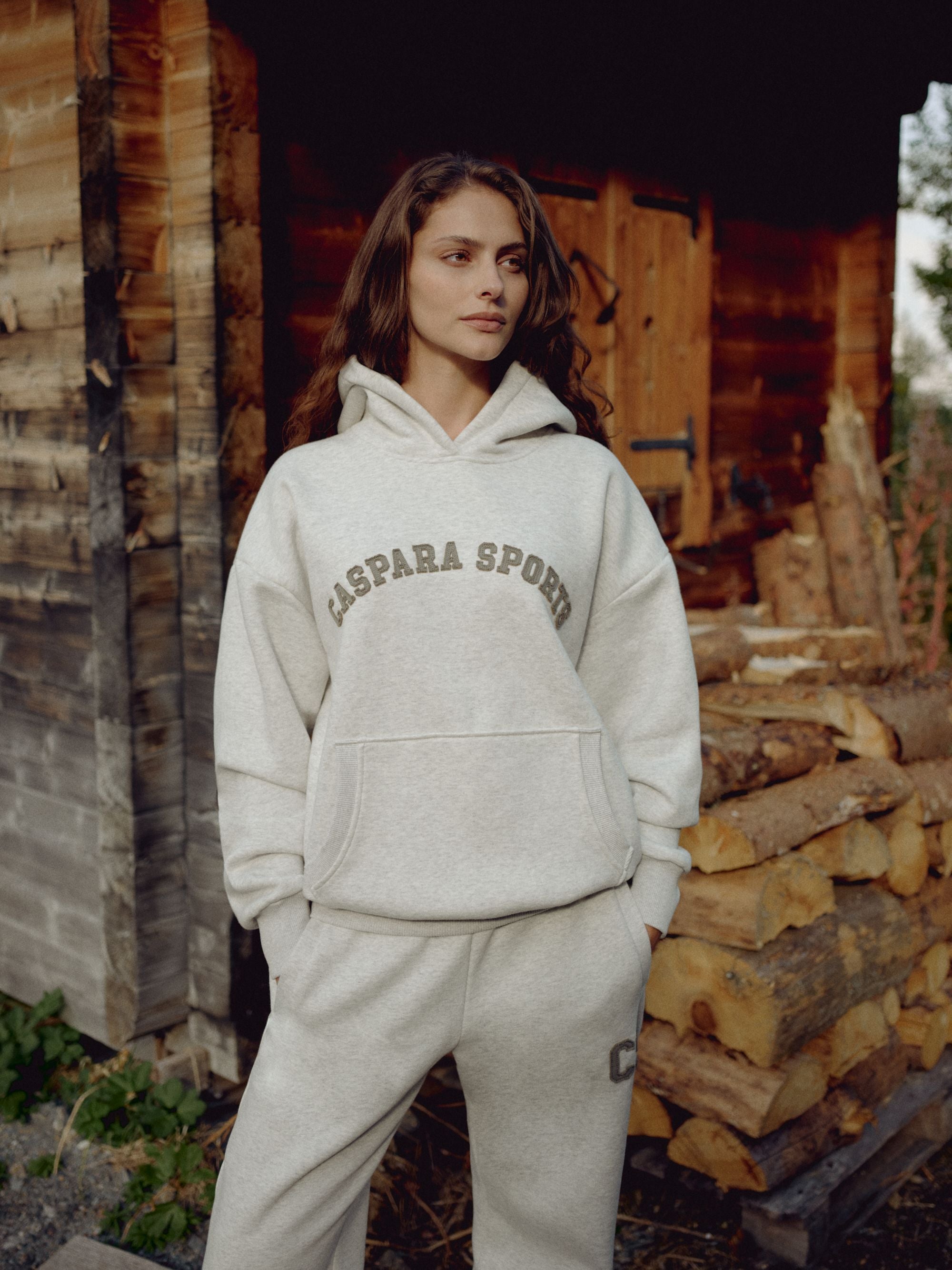 Woman wearing a grey melange CASPARA CS hoodie outside a wooden cabin.