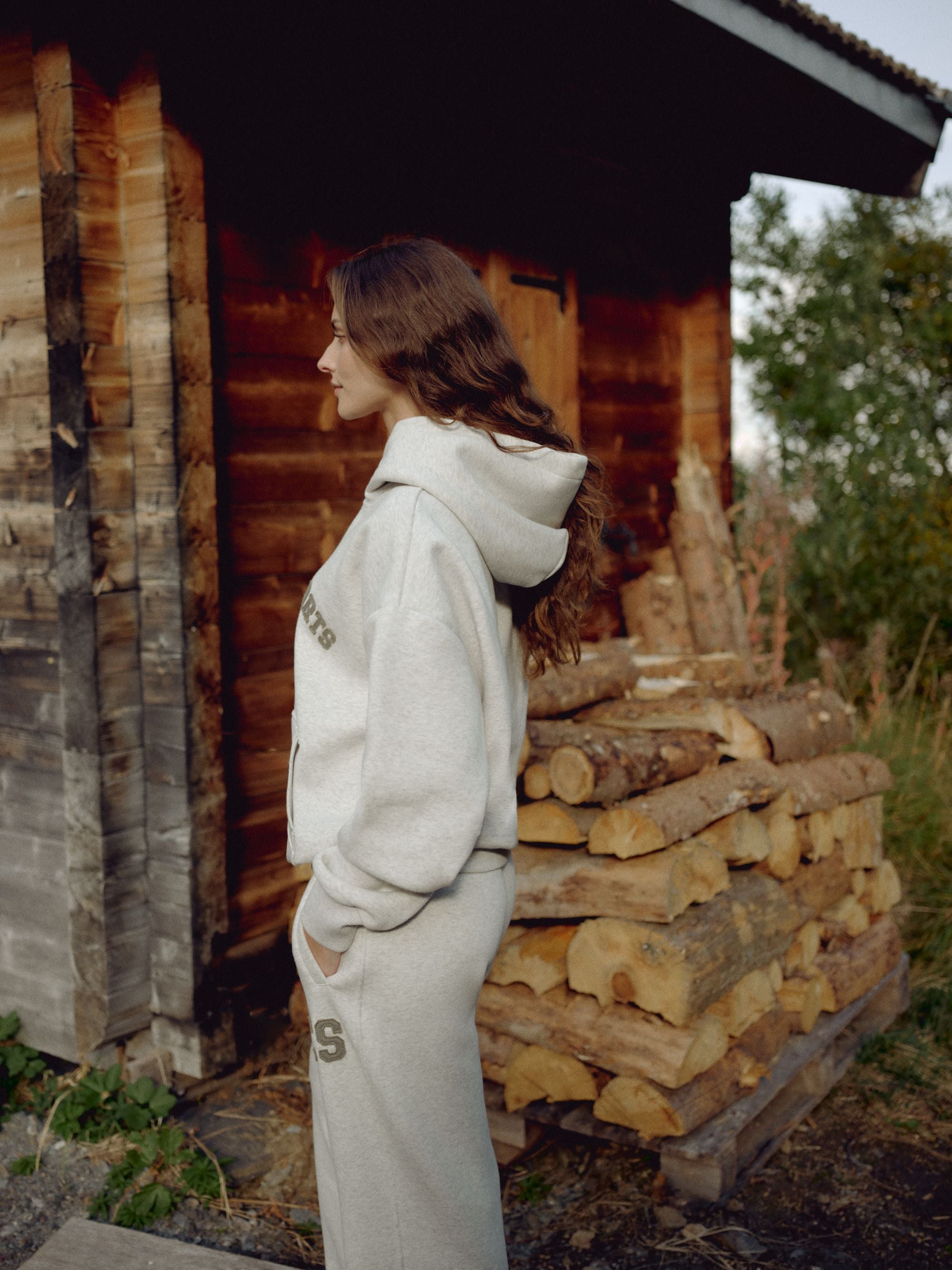Woman wearing a grey melange CASPARA CS hoodie next to stacked firewood outside a wooden cabin.