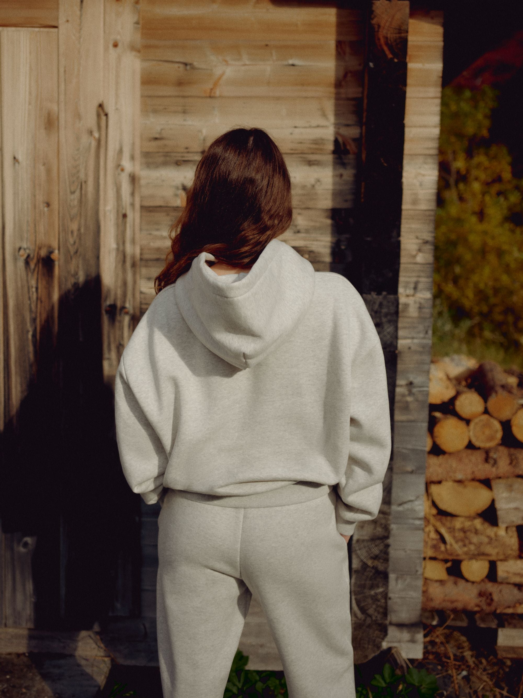 Woman wearing a grey melange CASPARA CS hoodie seen from behind beside a rustic wooden cabin.