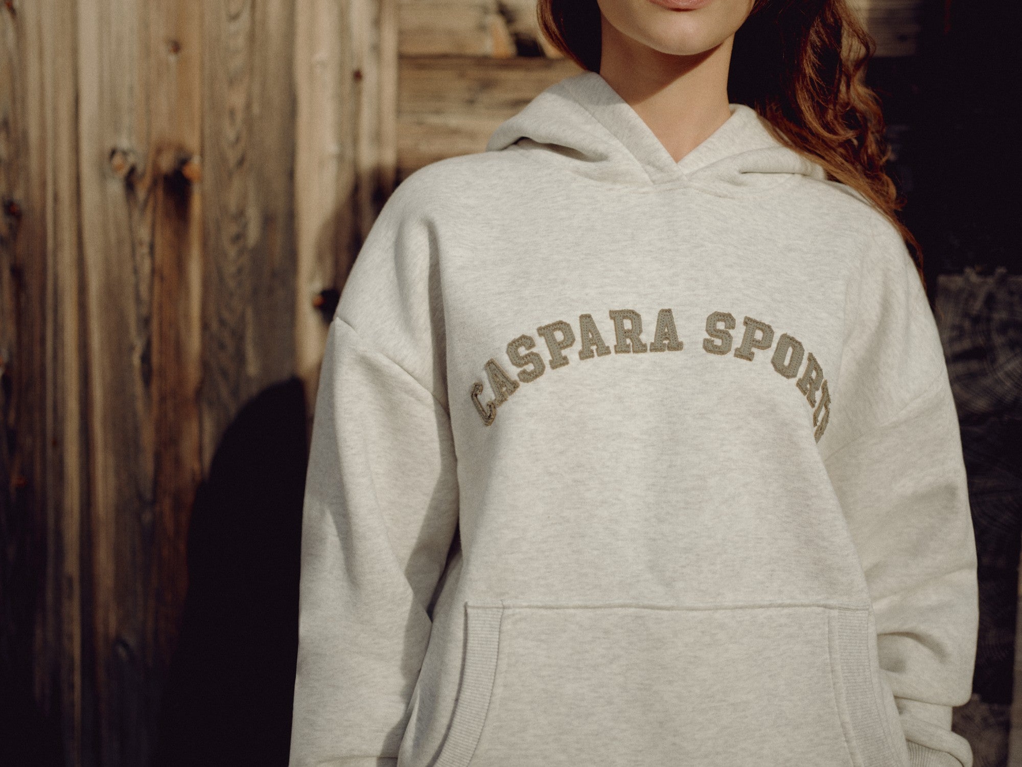 Woman wearing a grey melange CASPARA CS hoodie with khaki lettering against a wooden wall.