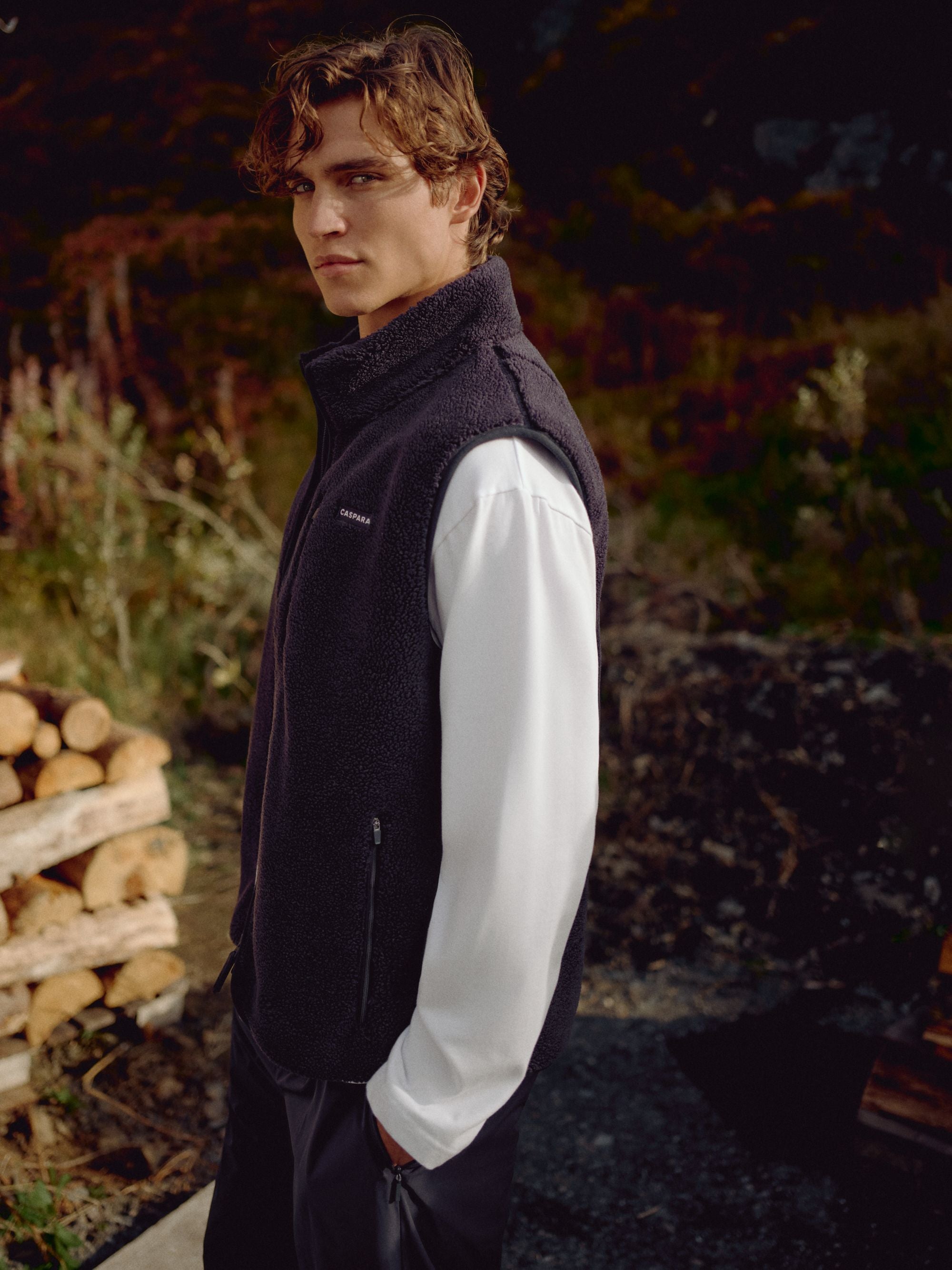 Man wearing a navy CASPARA Cloudfleece vest outdoors beside stacked firewood.