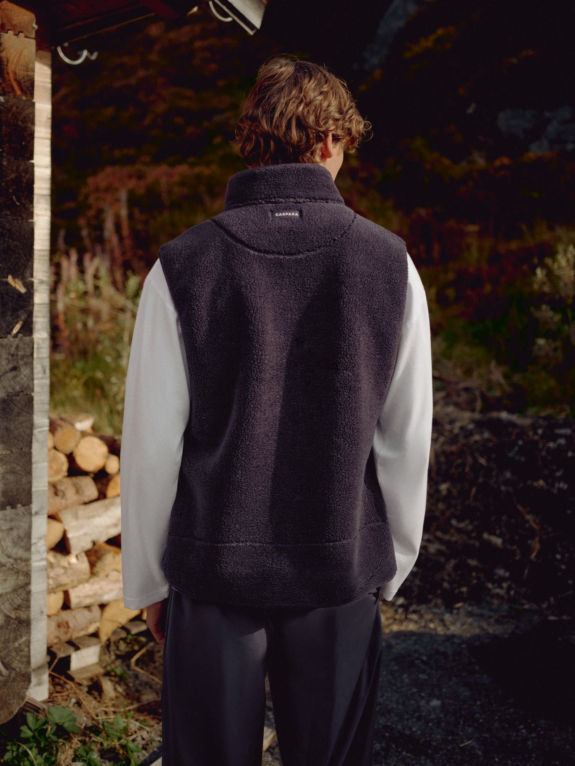 Back view of a man wearing a navy CASPARA Cloudfleece vest outdoors by stacked firewood.