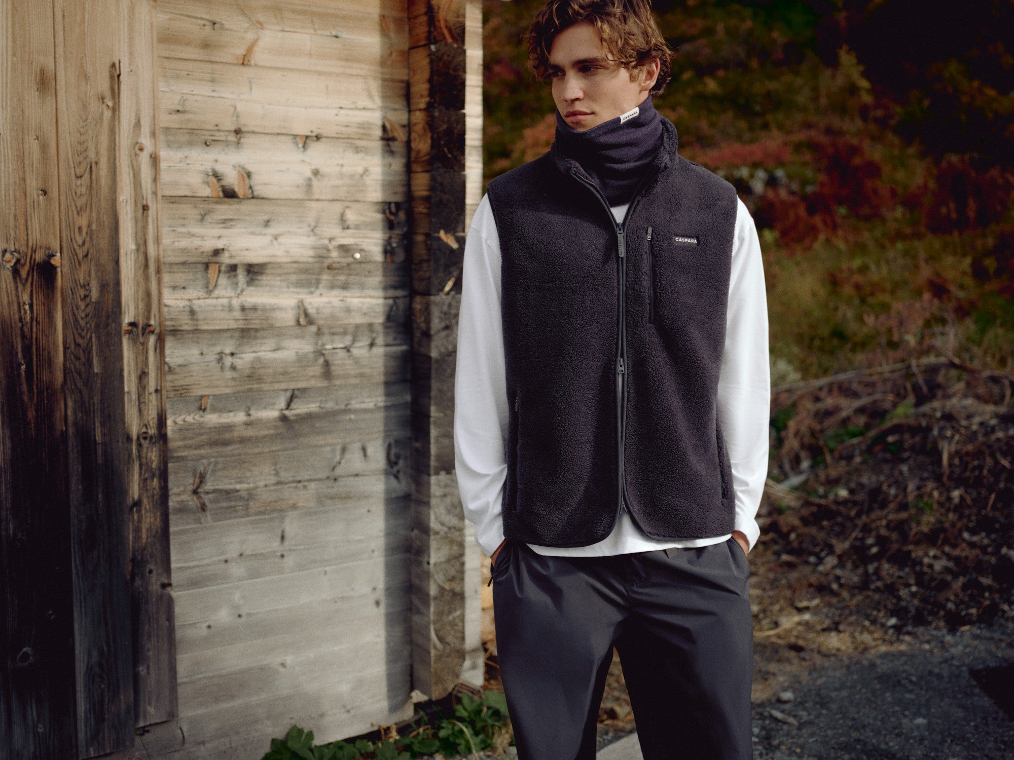 Man wearing a navy CASPARA Classic Neckwarmer and fleece vest outdoors by a wooden cabin.