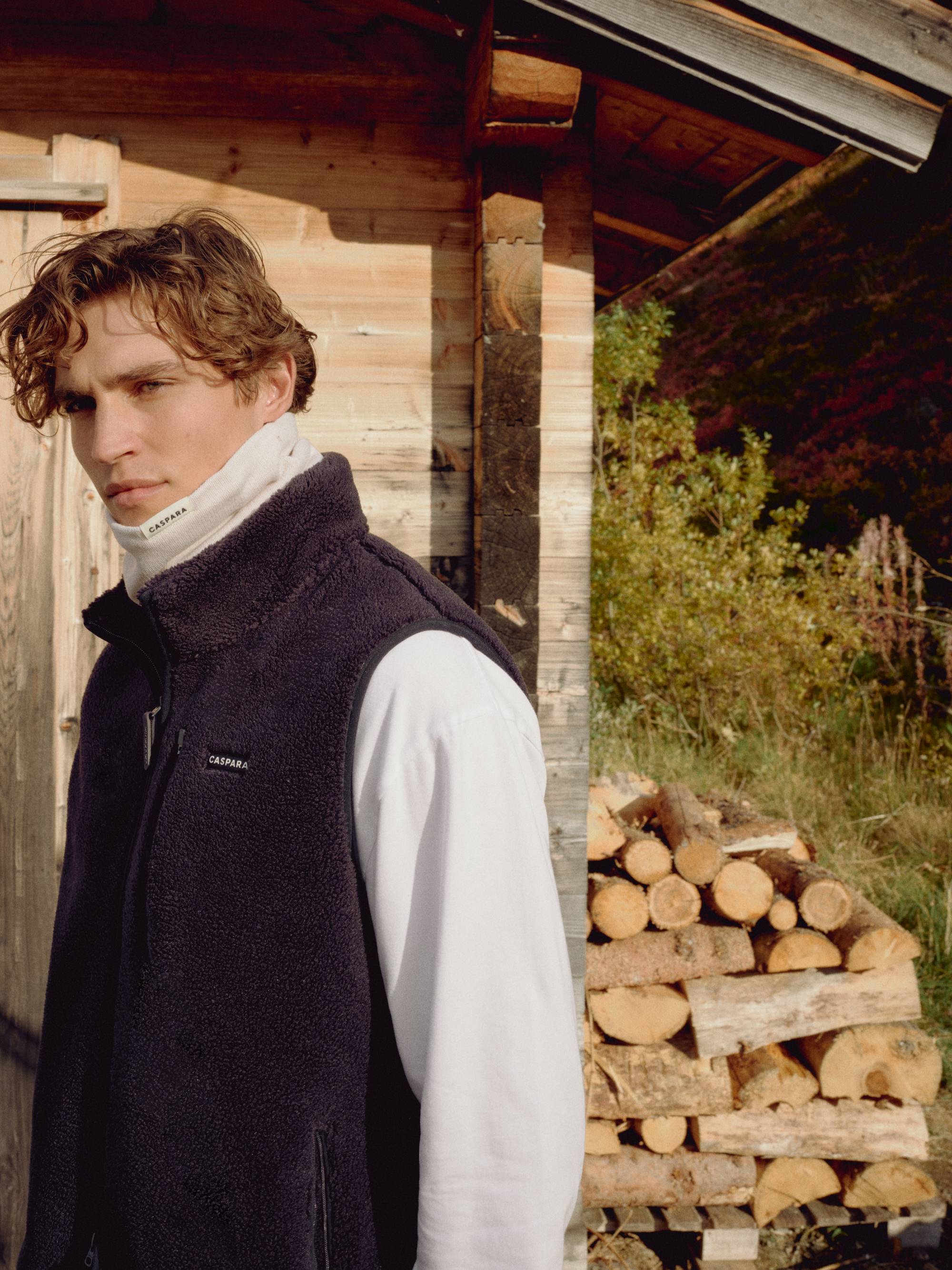 Man wearing a creme CASPARA Classic Neckwarmer outdoors by a wooden cabin.