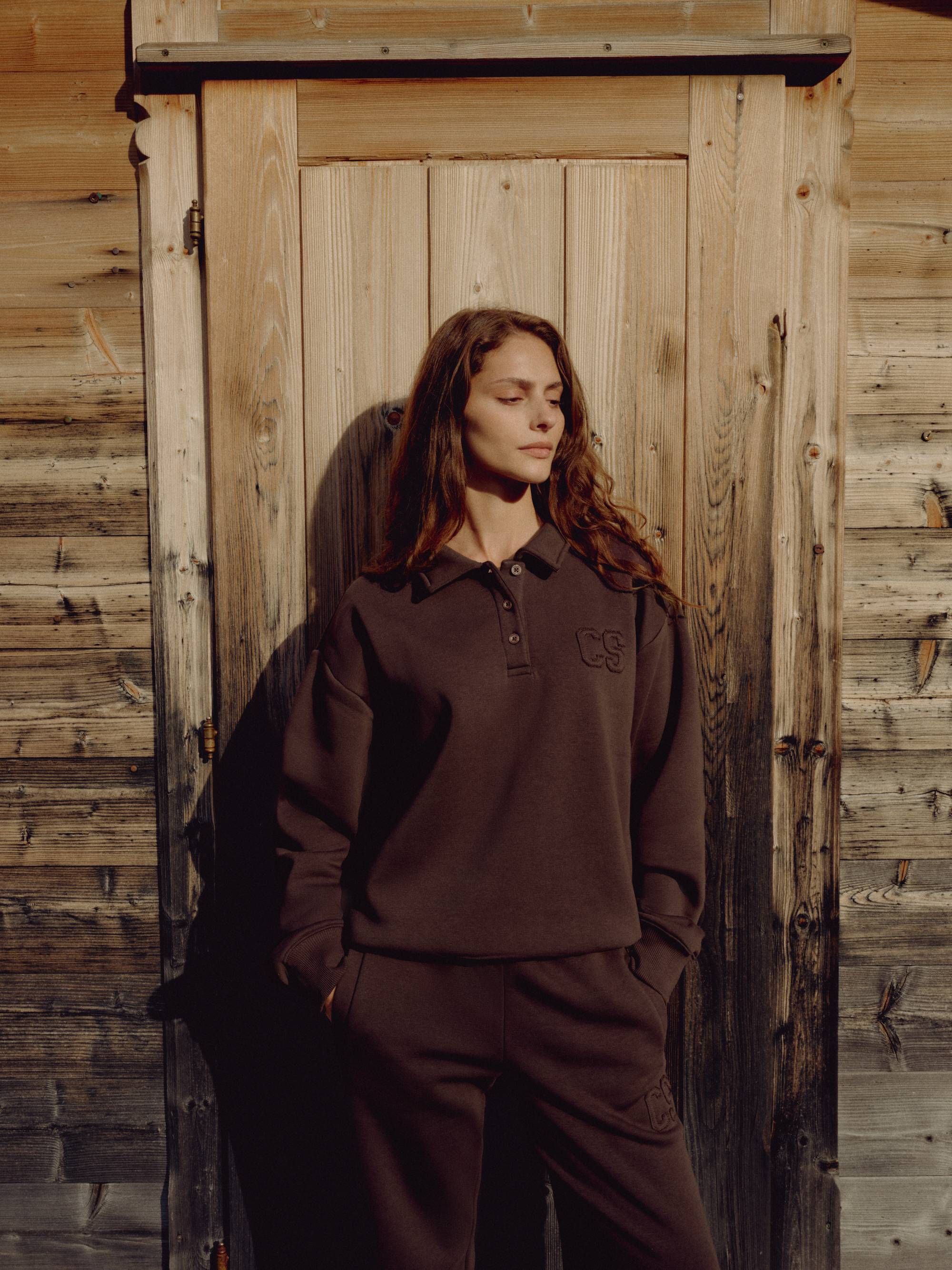 Woman wearing an espresso CASPARA CS rugby shirt standing in front of a rustic wooden door.