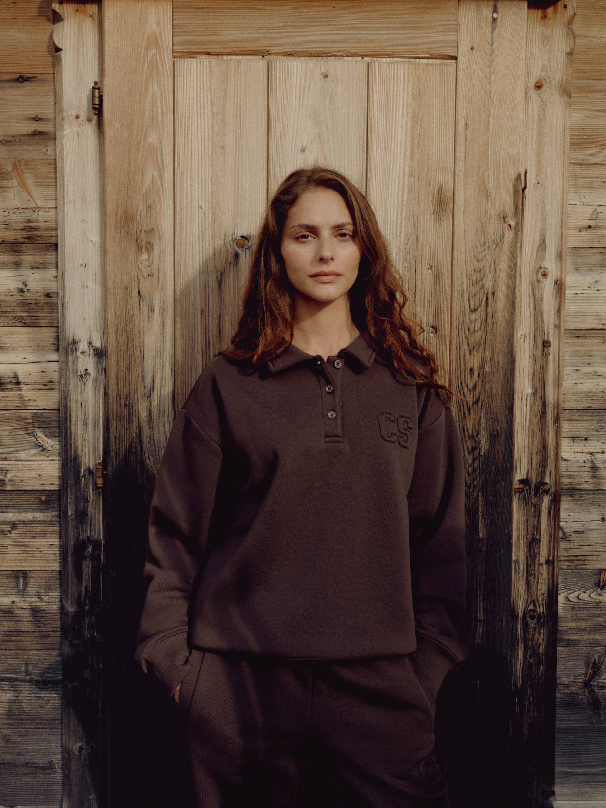 Woman wearing an espresso CASPARA CS rugby shirt standing in front of a wooden door.