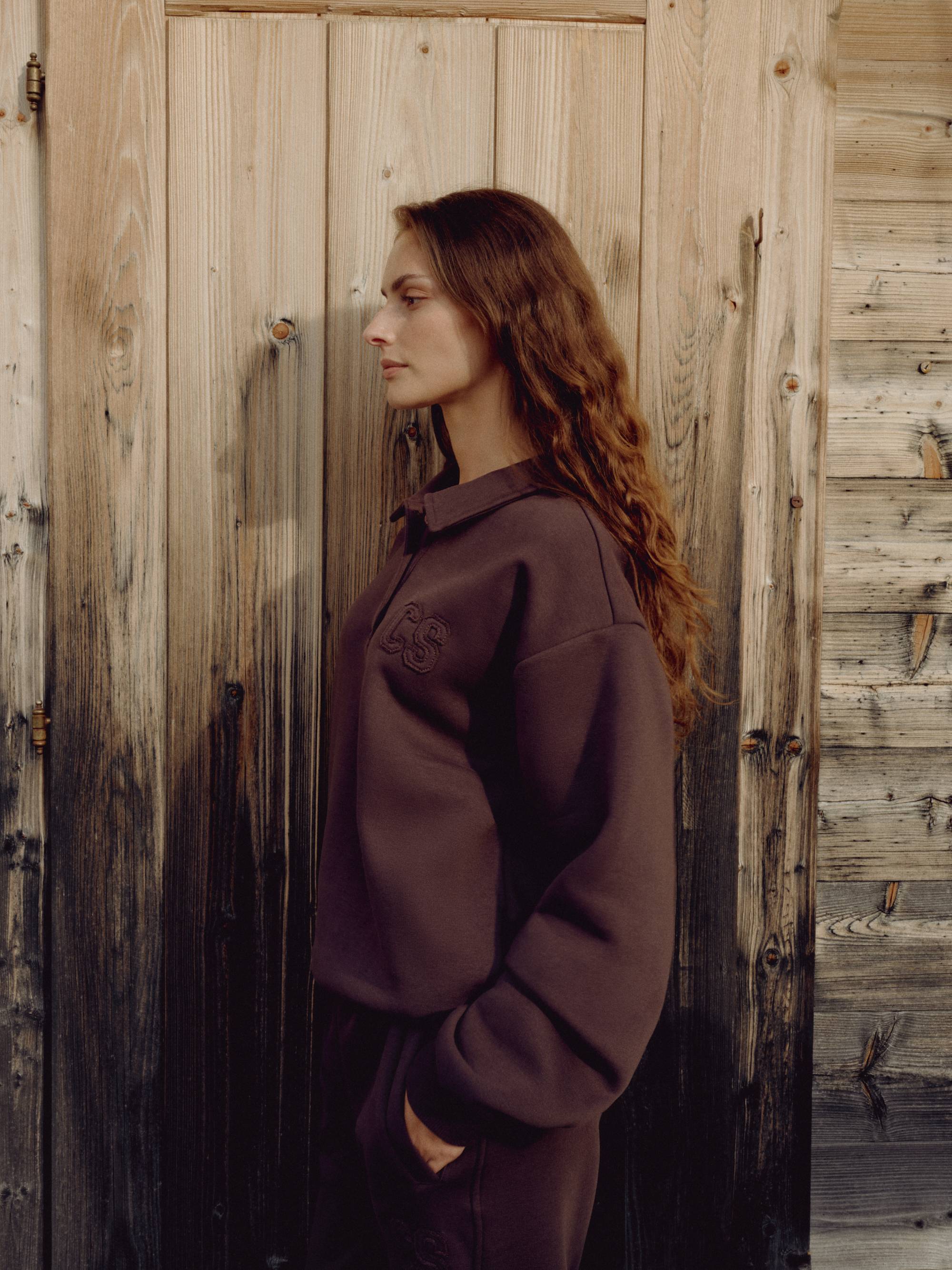Woman wearing an espresso CASPARA CS rugby shirt standing against a wooden wall.