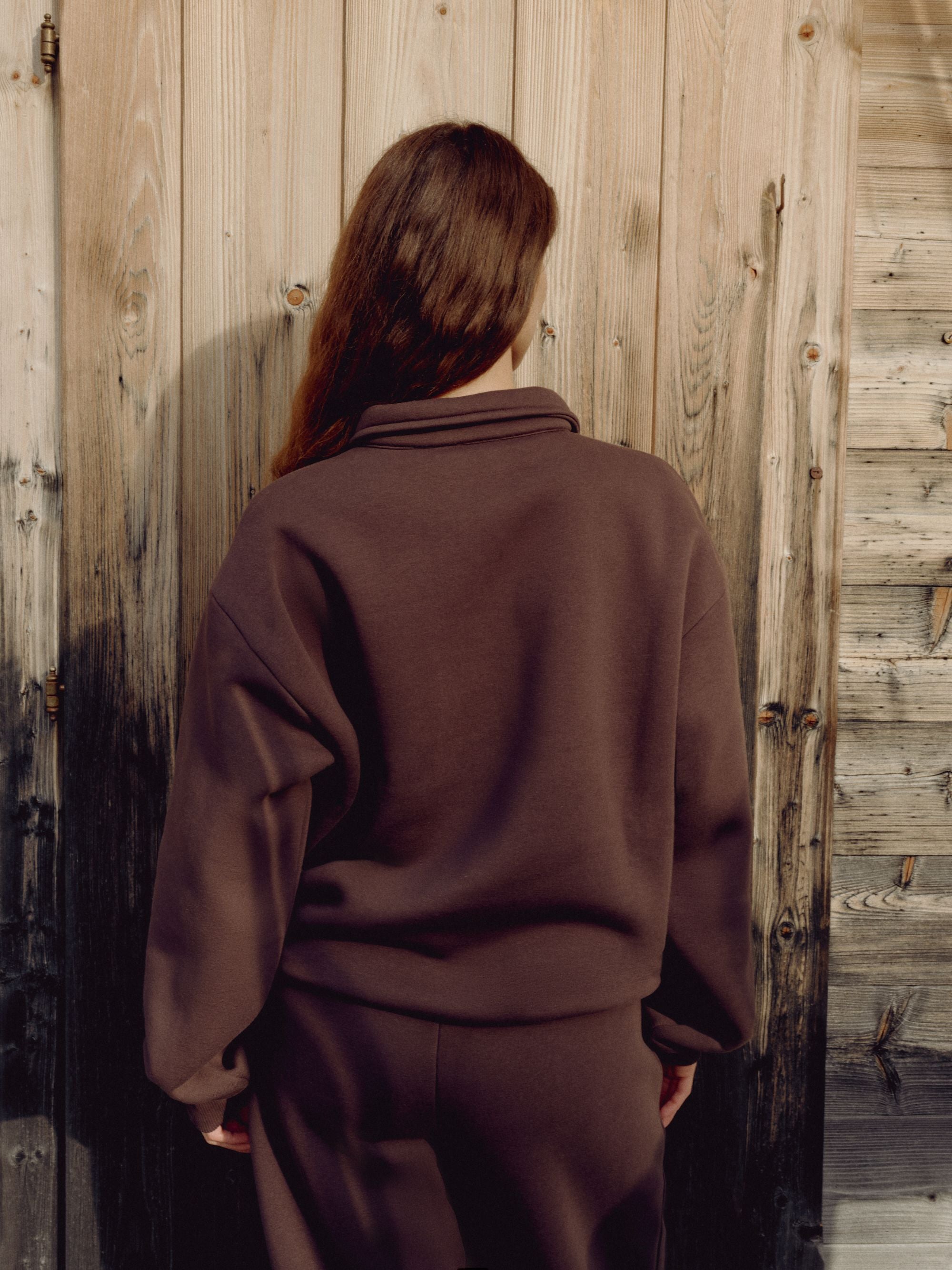Woman wearing an espresso CASPARA CS rugby shirt standing in front of a wooden wall.