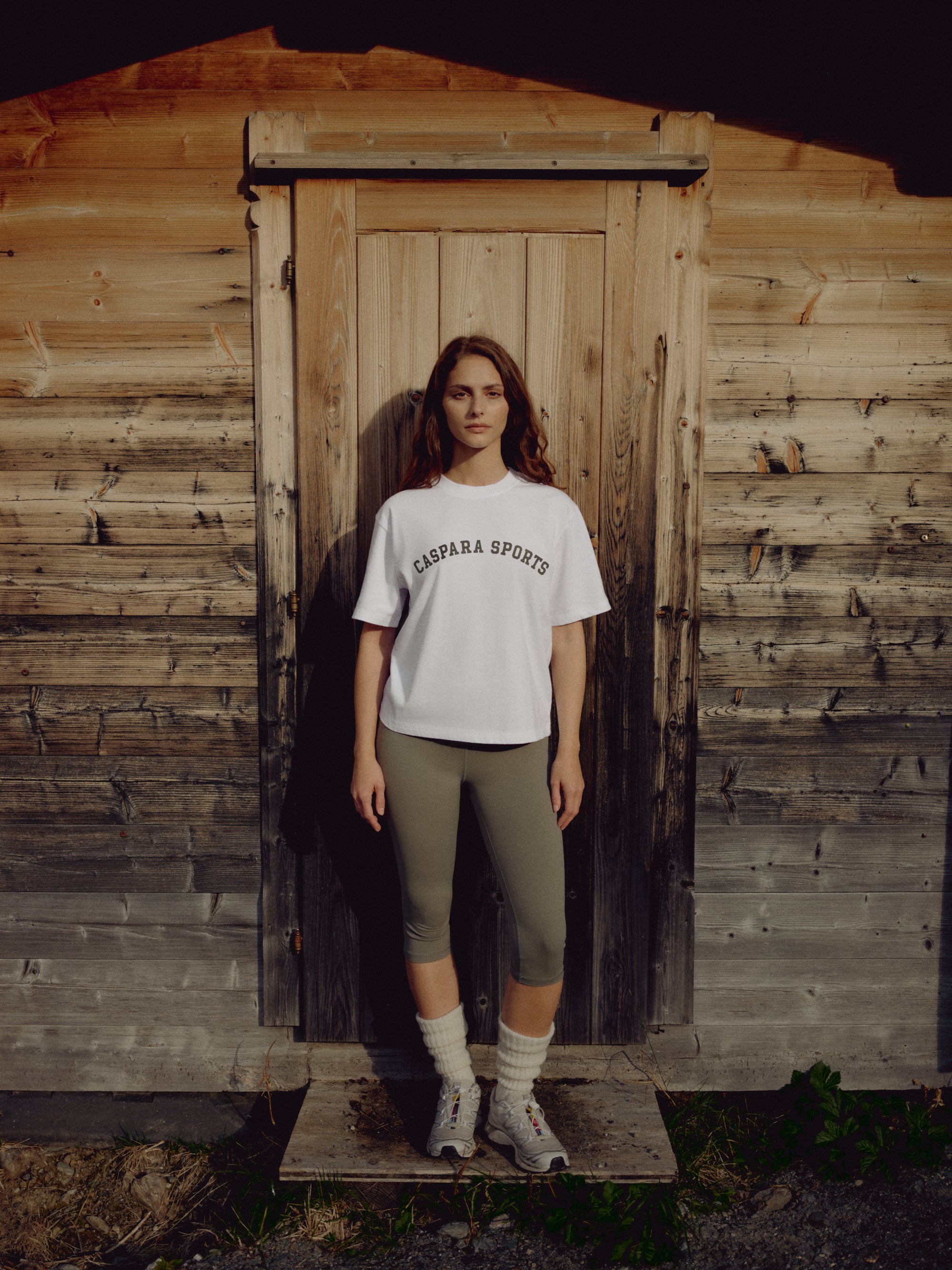 Woman wearing a white CASPARA CS Signature tee standing in front of a rustic wooden door.