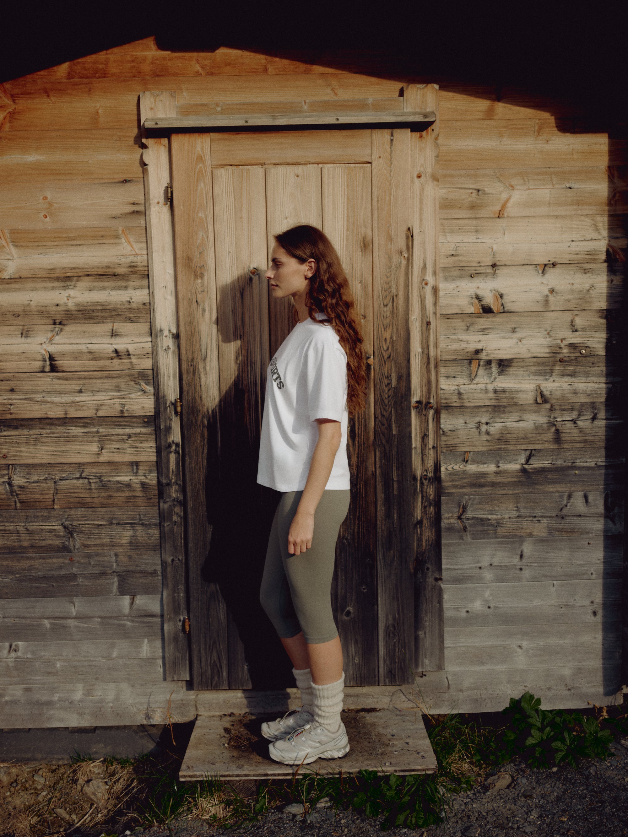Woman wearing a white CASPARA CS Signature tee standing in front of a wooden cabin.