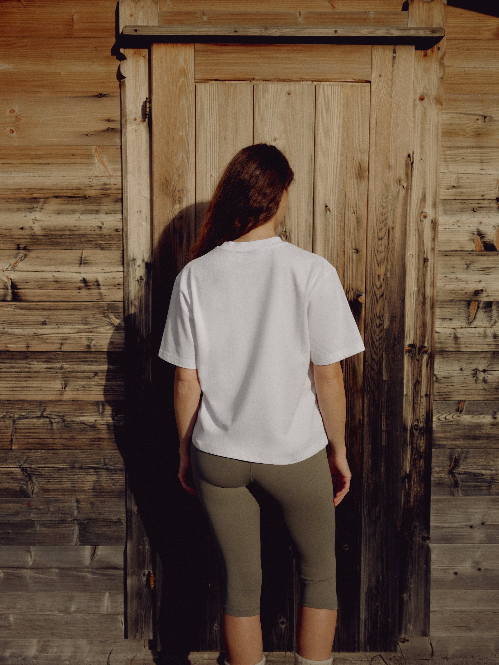 Woman seen from behind wearing a white CASPARA CS Signature tee against a rustic wooden door.