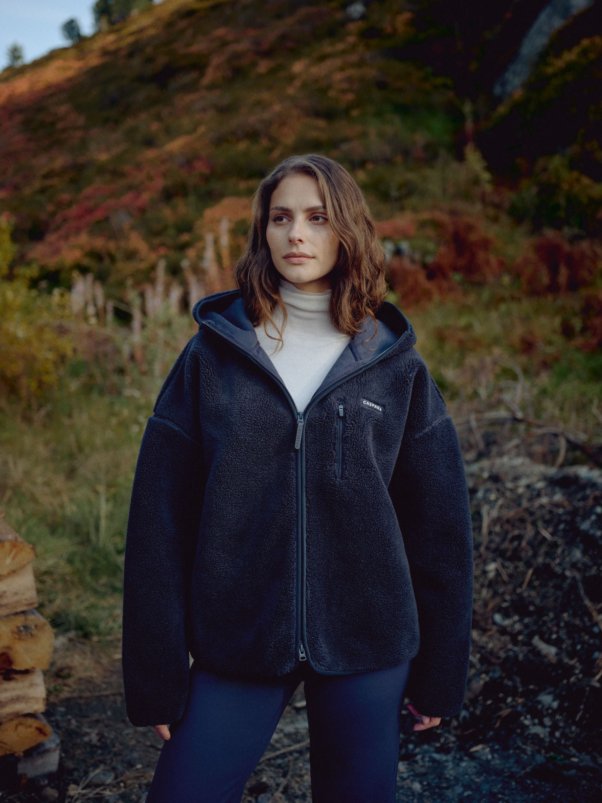 Woman wearing a navy CASPARA Cloudfleece hooded jacket on an autumn hillside.