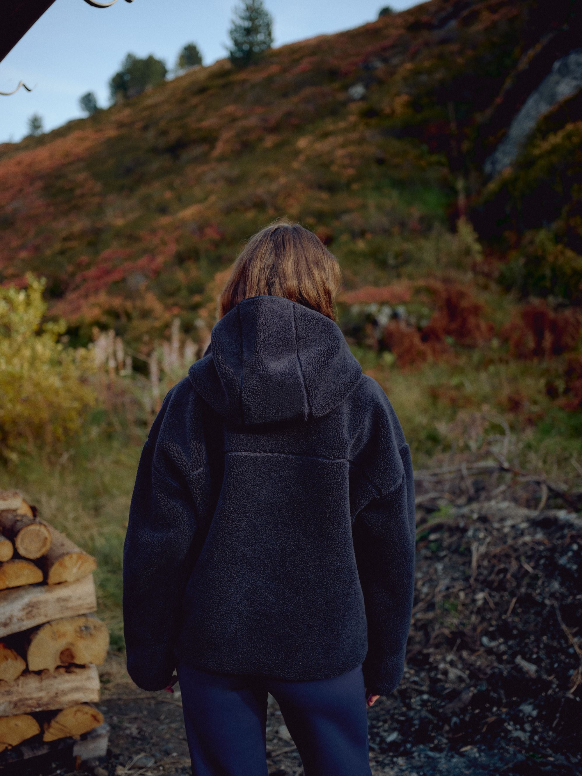 Woman wearing a navy CASPARA Cloudfleece hooded jacket, viewed from behind outdoors.
