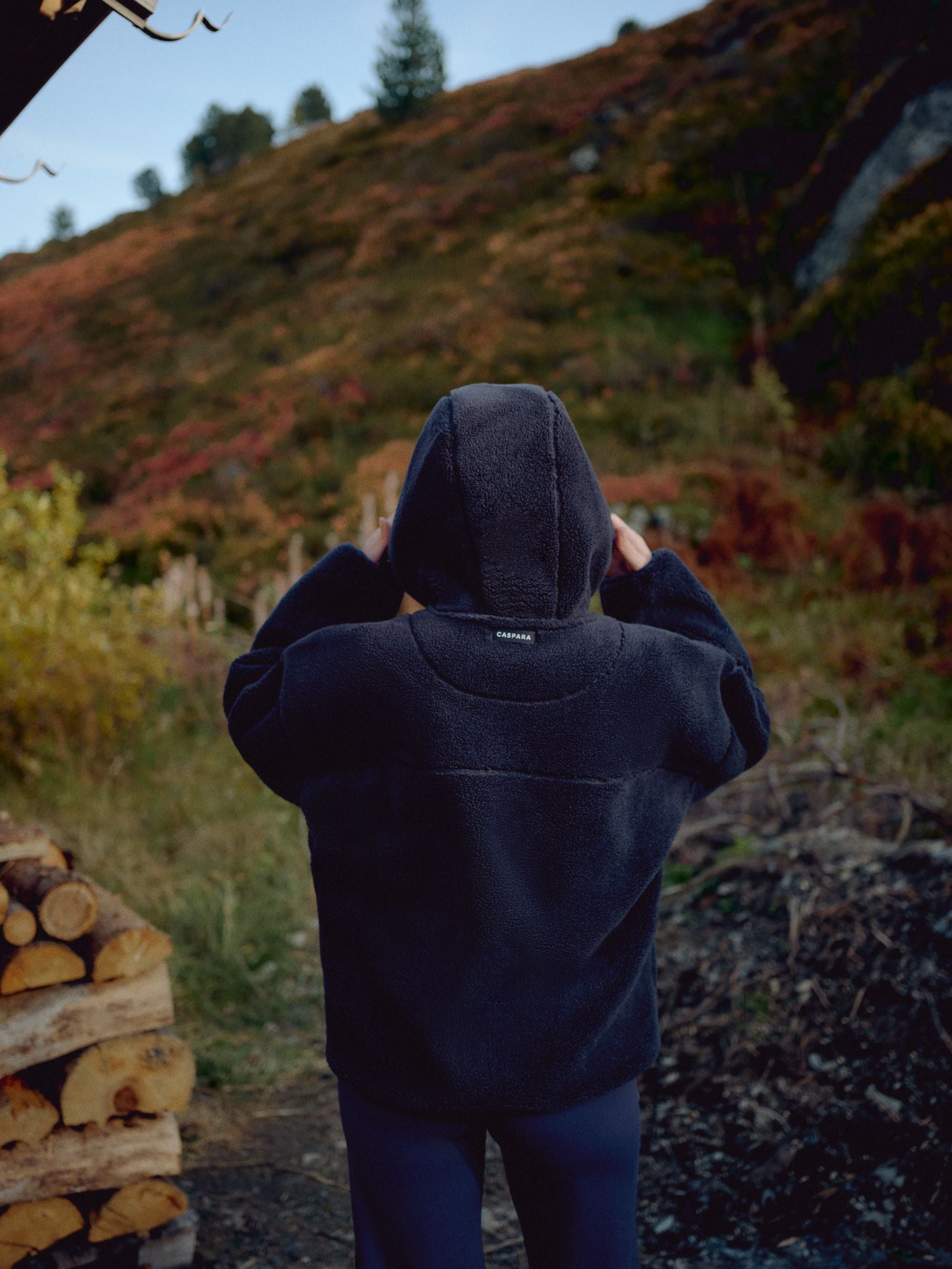 Back view of a woman wearing a navy CASPARA Cloudfleece hooded jacket on a hillside.
