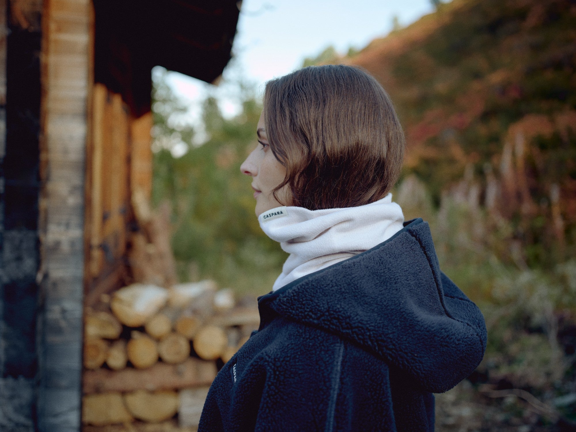 Woman wearing a creme CASPARA Classic Neckwarmer outdoors next to a wooden cabin.