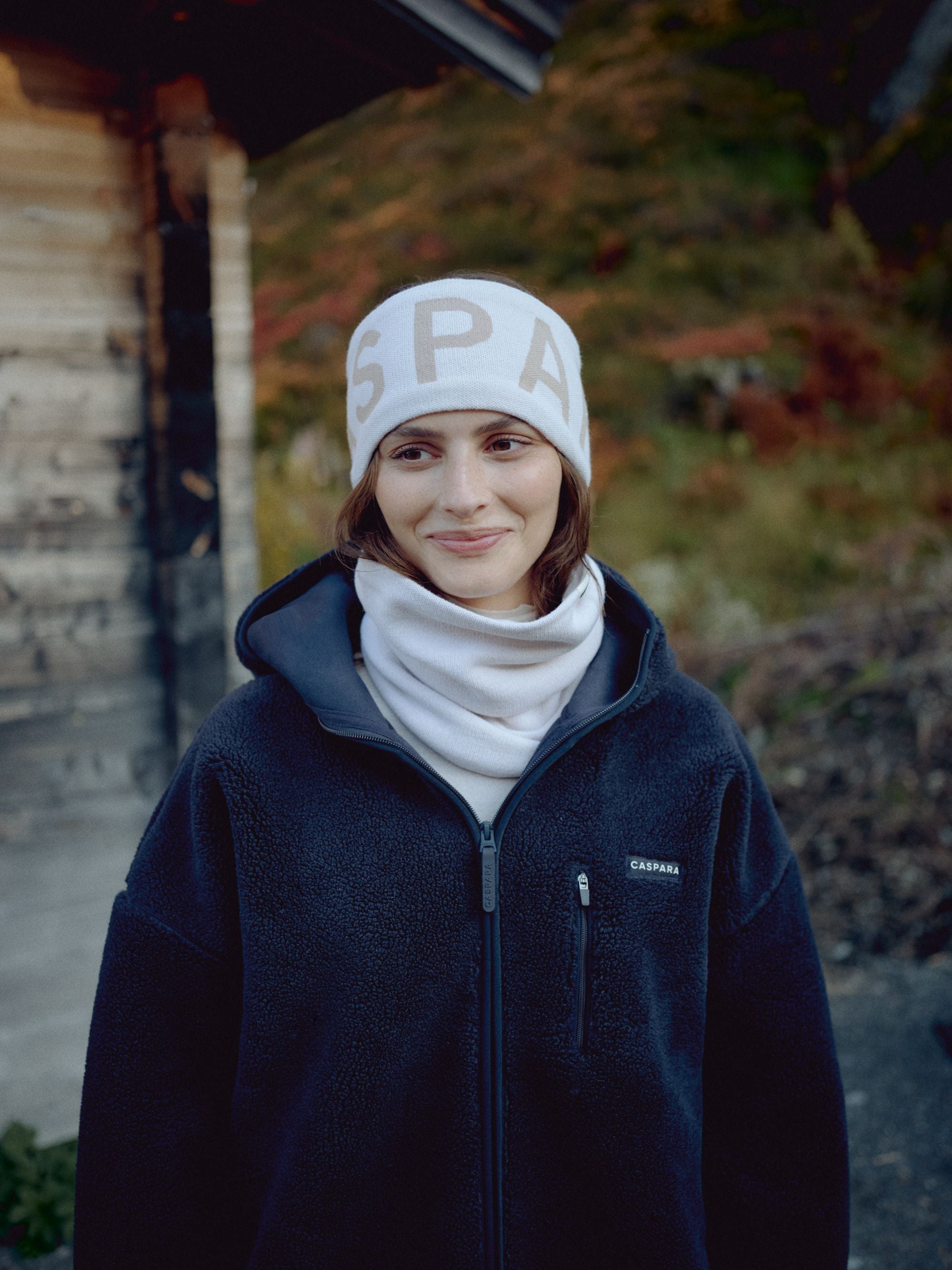 Woman wearing a creme CASPARA SKI headband outdoors by a wooden cabin.