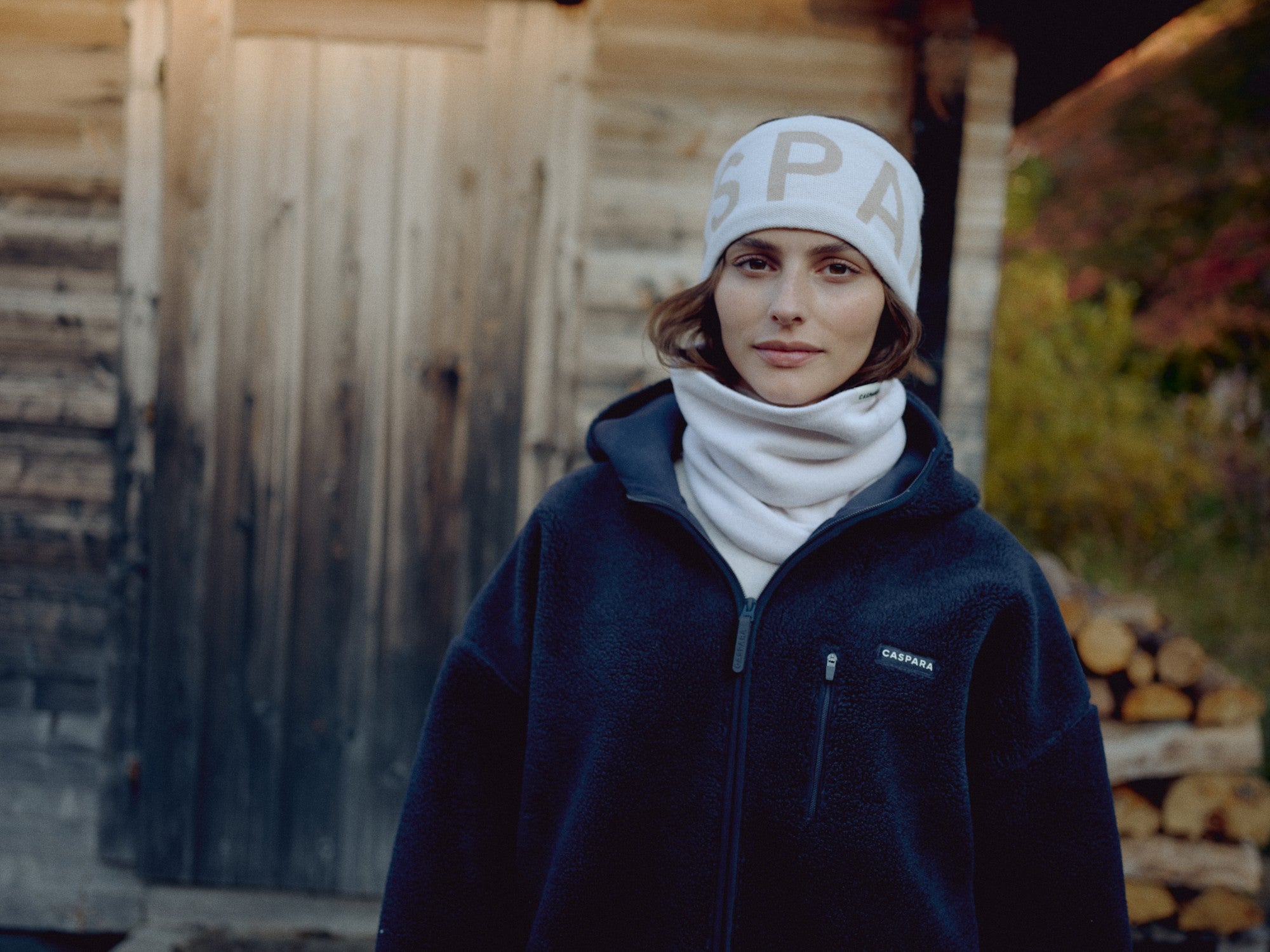 Woman wearing a creme CASPARA SKI headband in front of a rustic cabin.