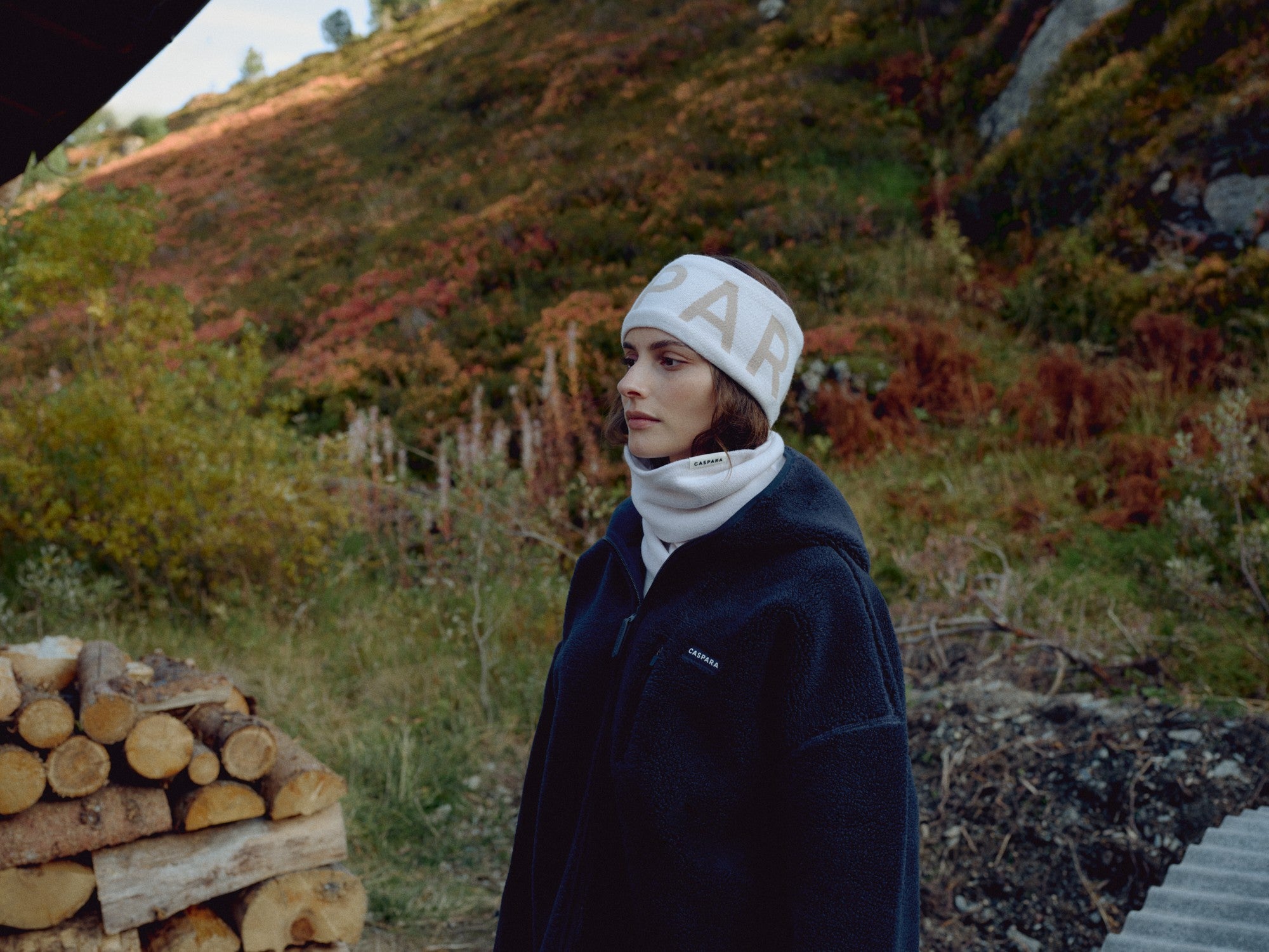 Woman wearing a creme CASPARA SKI headband outdoors near stacked logs.