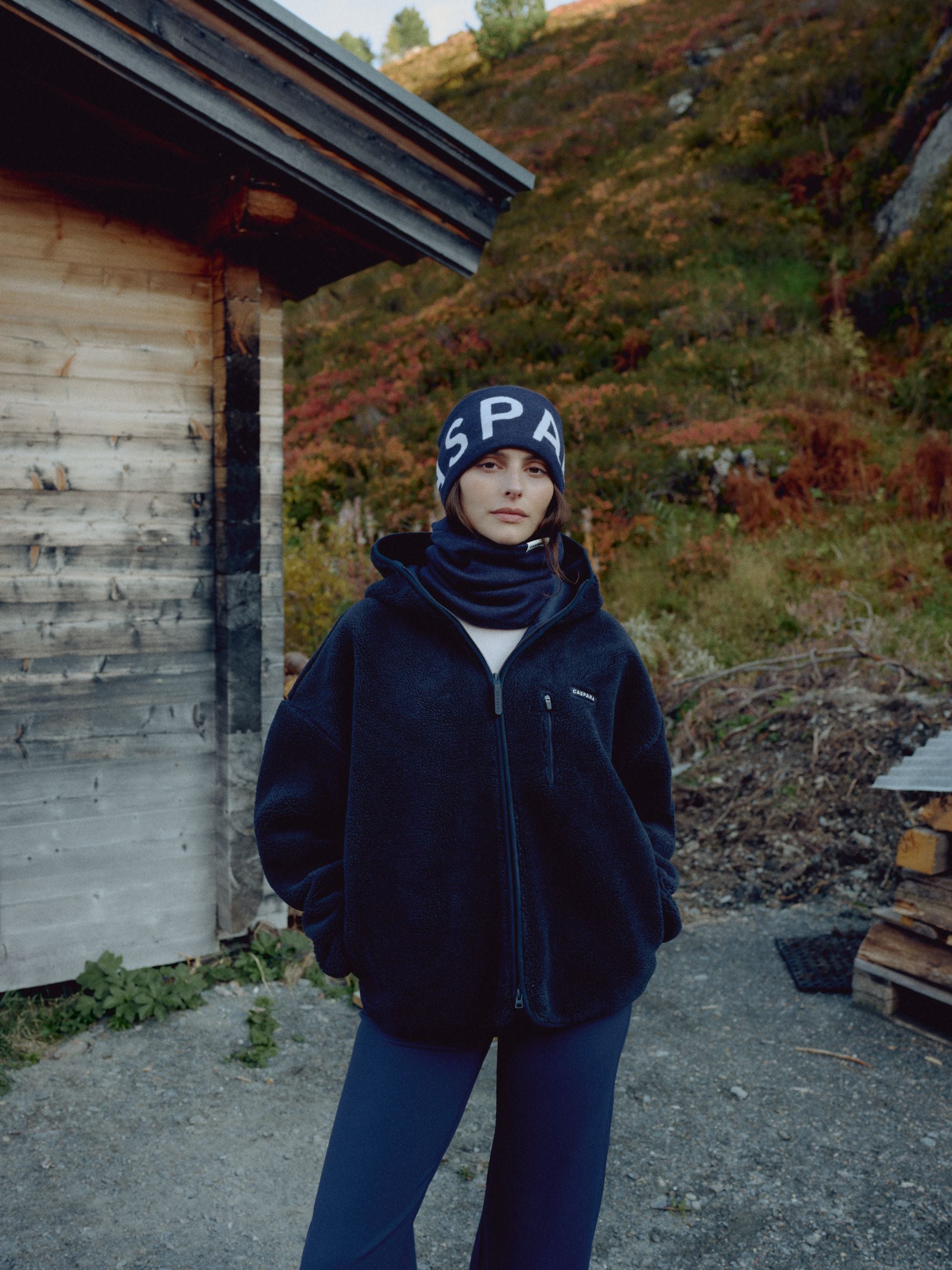 Woman wearing a navy CASPARA SKI headband and fleece jacket outside a wooden mountain cabin.