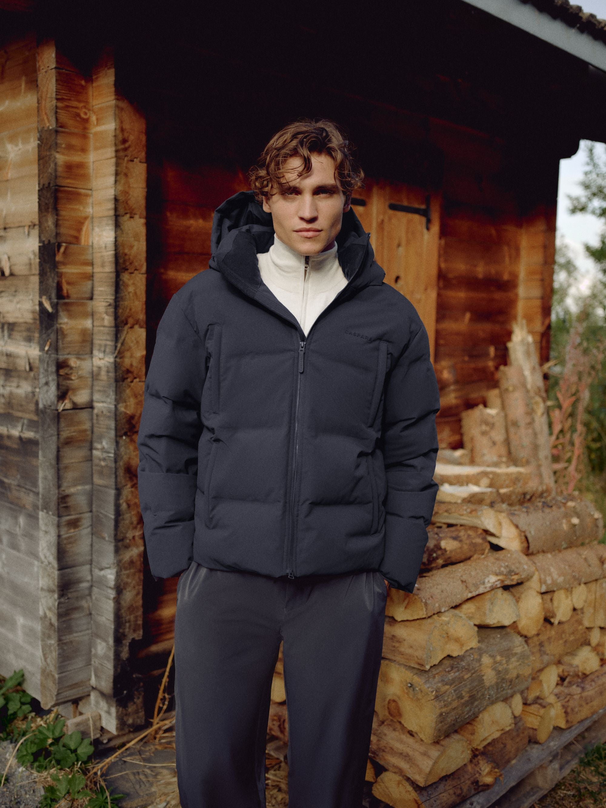 Man wearing a navy CASPARA Boreal puffer jacket standing outside a wooden cabin.