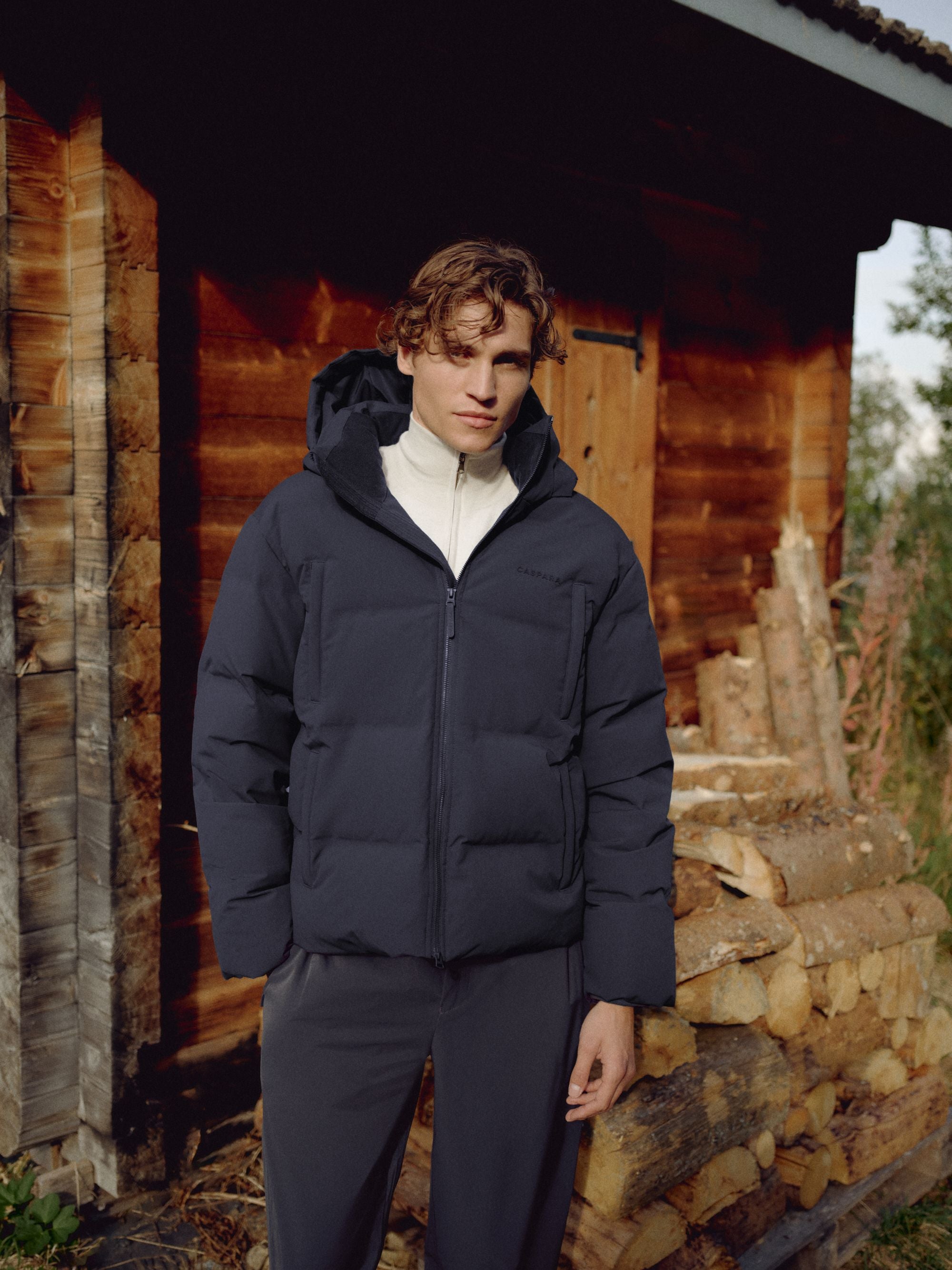 Man wearing a navy CASPARA Boreal puffer jacket standing outside a wooden cabin.