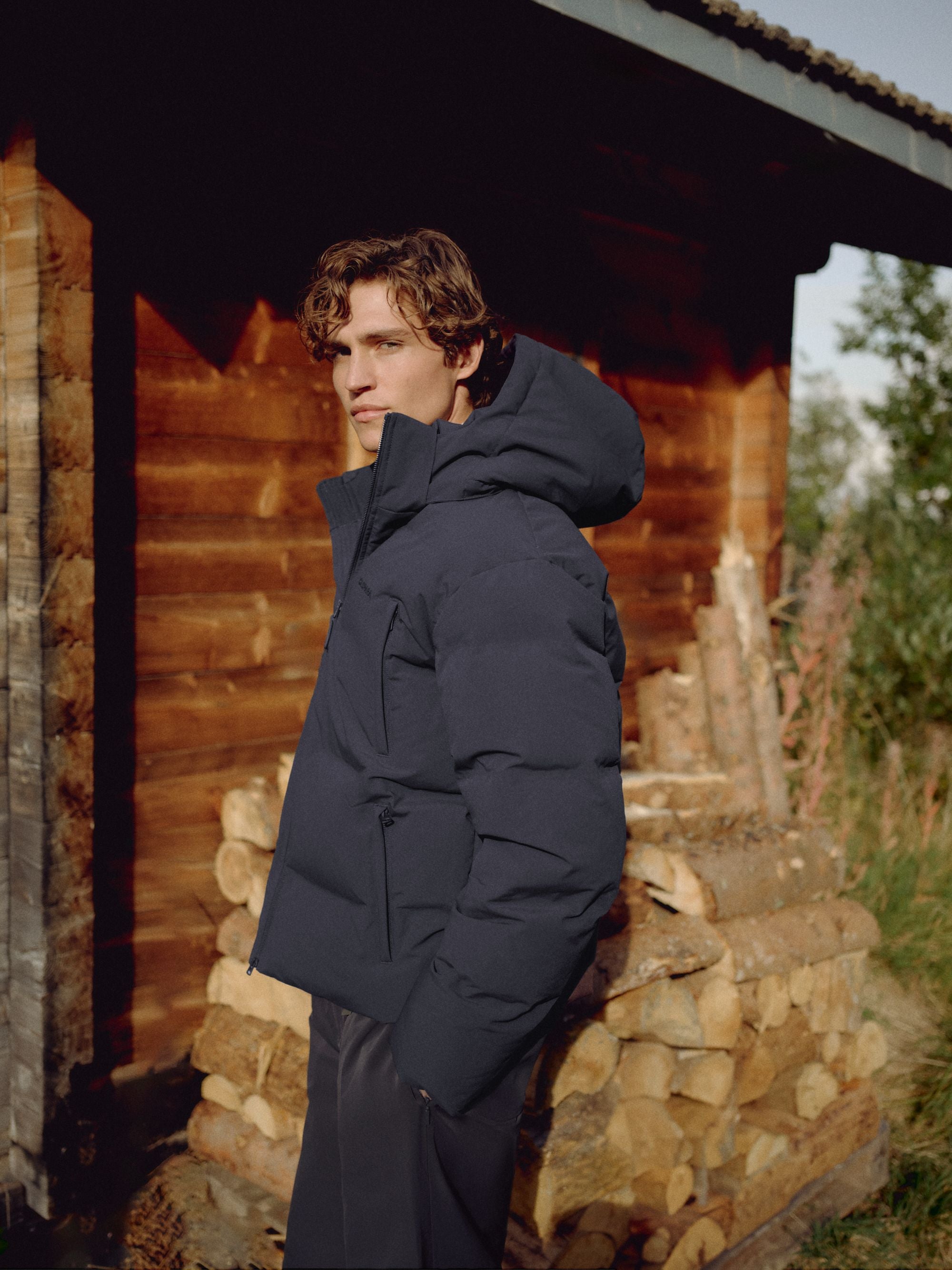 Man wearing a navy CASPARA Boreal puffer jacket standing outdoors next to stacked firewood.