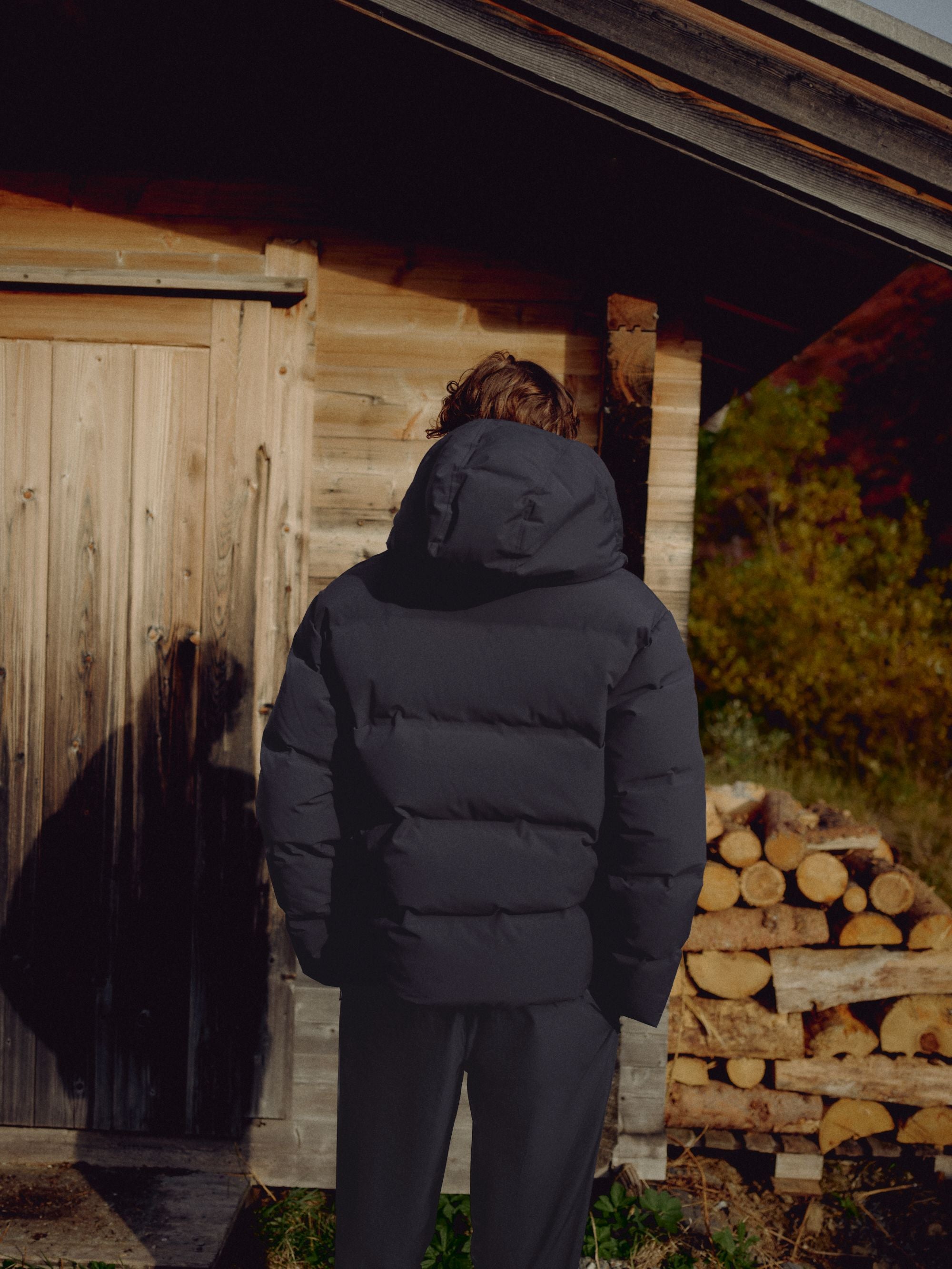 Man wearing a hooded navy CASPARA Boreal puffer jacket standing outside a wooden cabin.