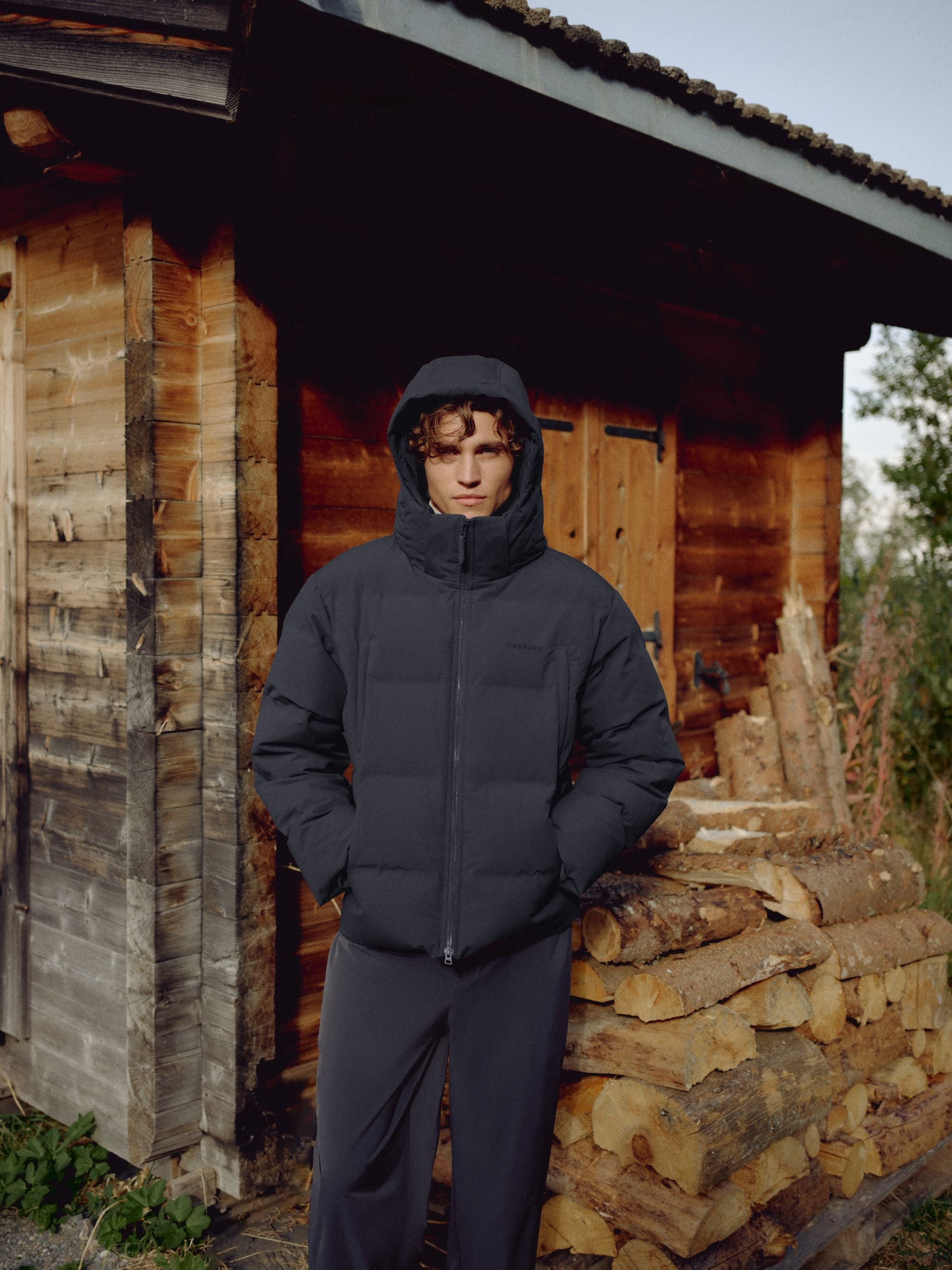 Man wearing a navy CASPARA Boreal puffer jacket with hood up outside a rustic wooden cabin.