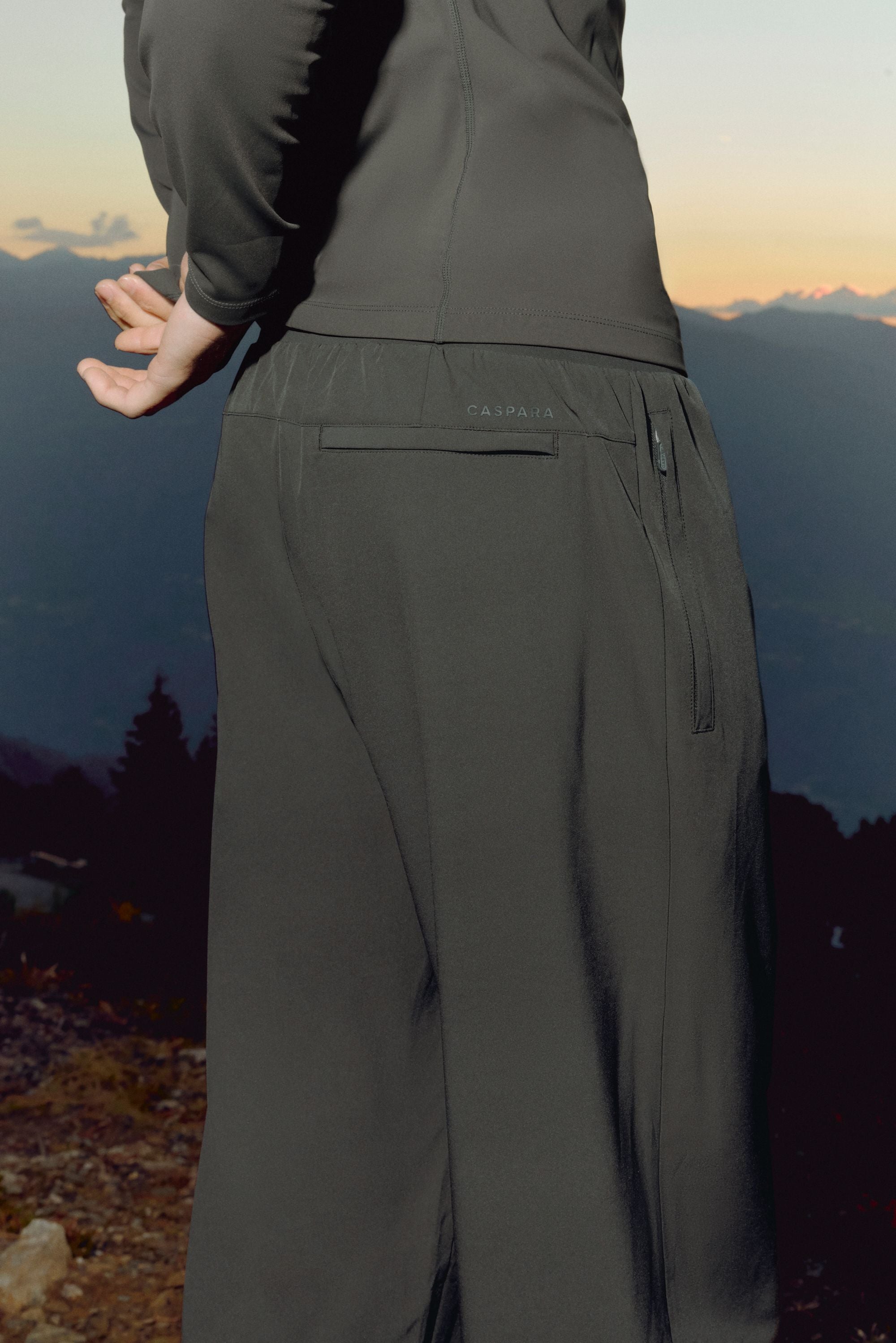Close-up rear view of olive green CASPARA Prime Core pants against a mountain landscape.