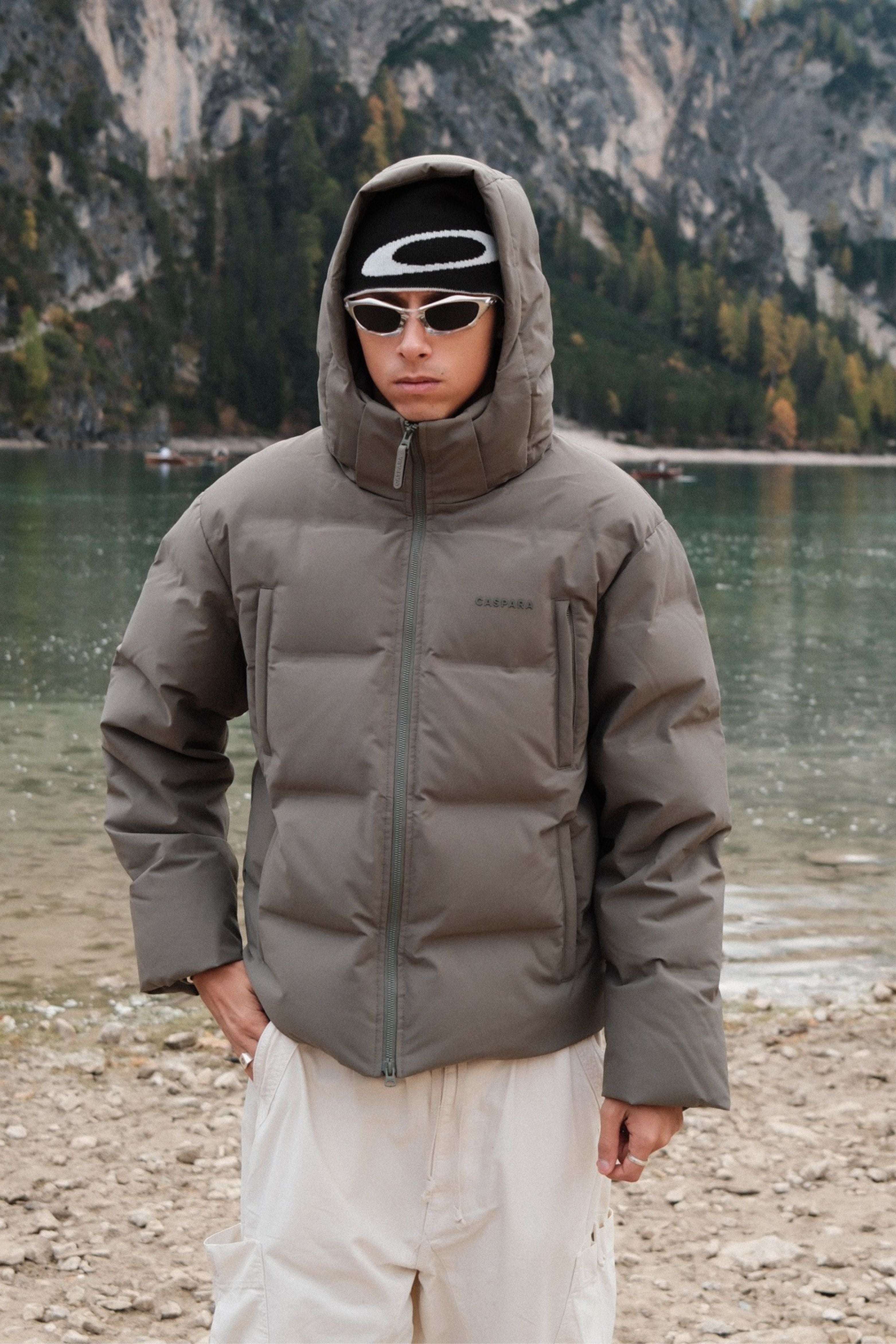 Man wearing a khaki CASPARA Boreal puffer jacket with the hood up by a mountain lake.