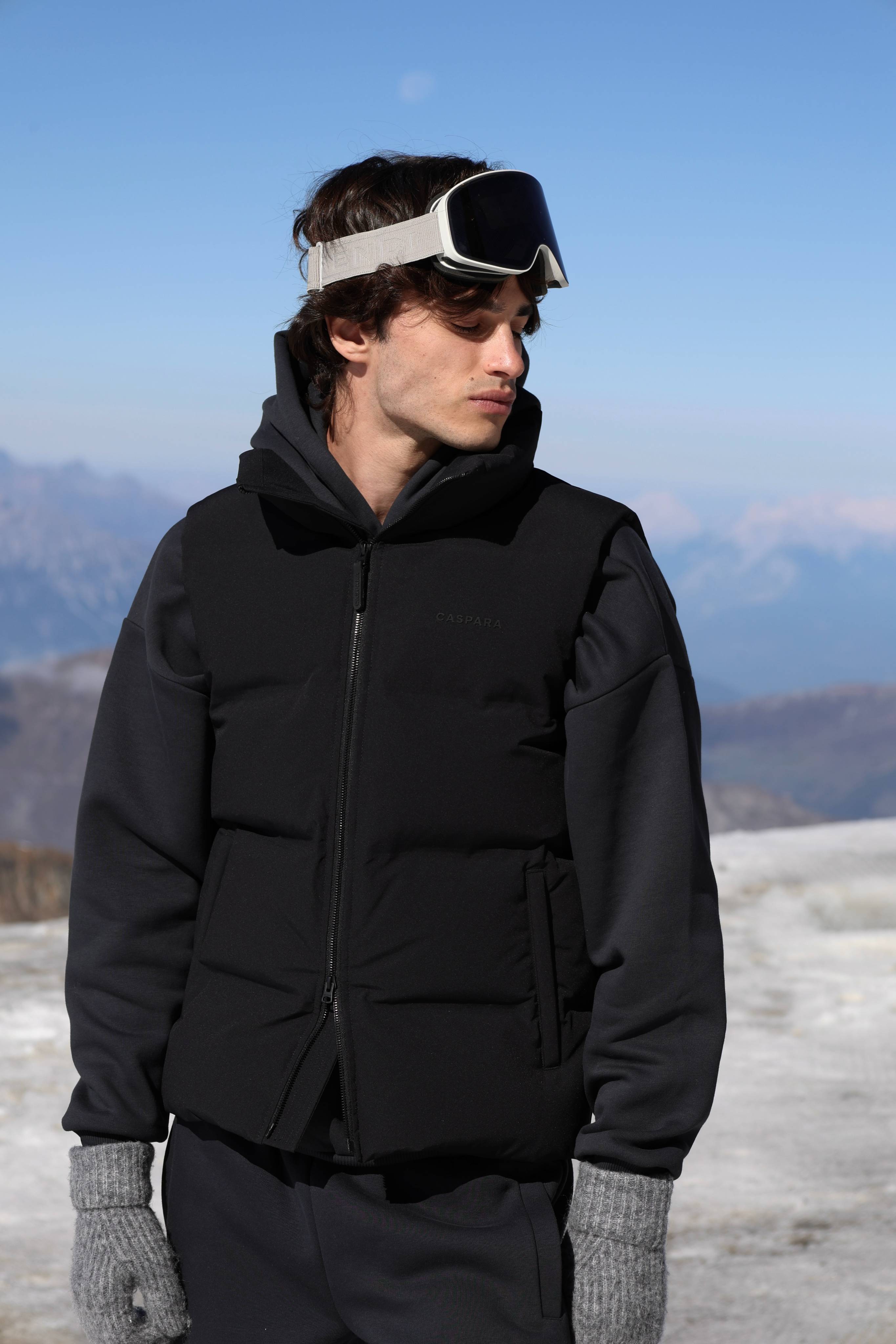 Man wearing a black CASPARA Boreal puffer vest outdoors in a snowy mountain setting.