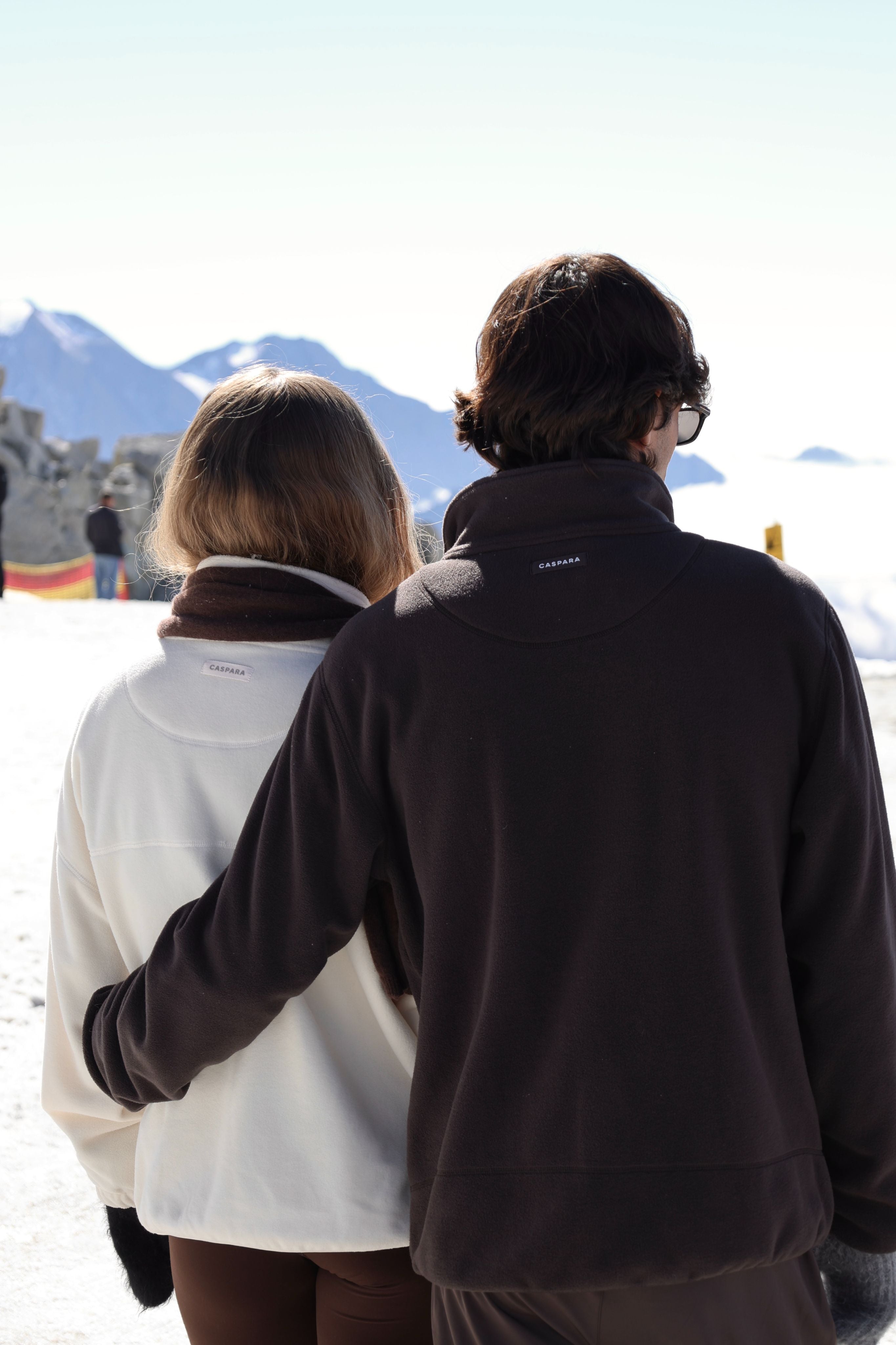 Back view of a couple wearing cocoa bean and creme CASPARA polar fleece pullovers on a snowy mountain.