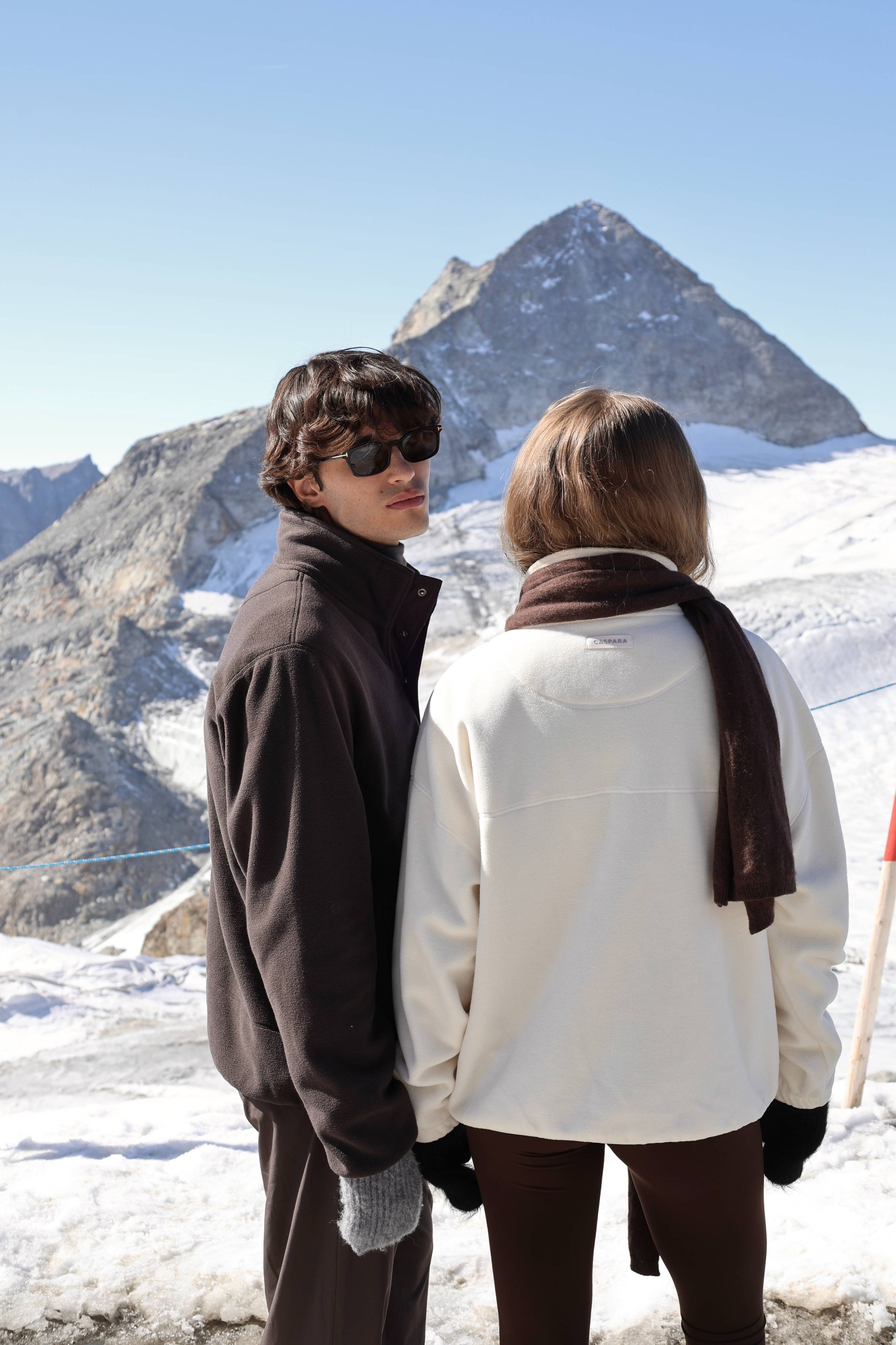Man wearing a cocoa bean CASPARA polar fleece pullover and woman in cream on a snowy mountain.